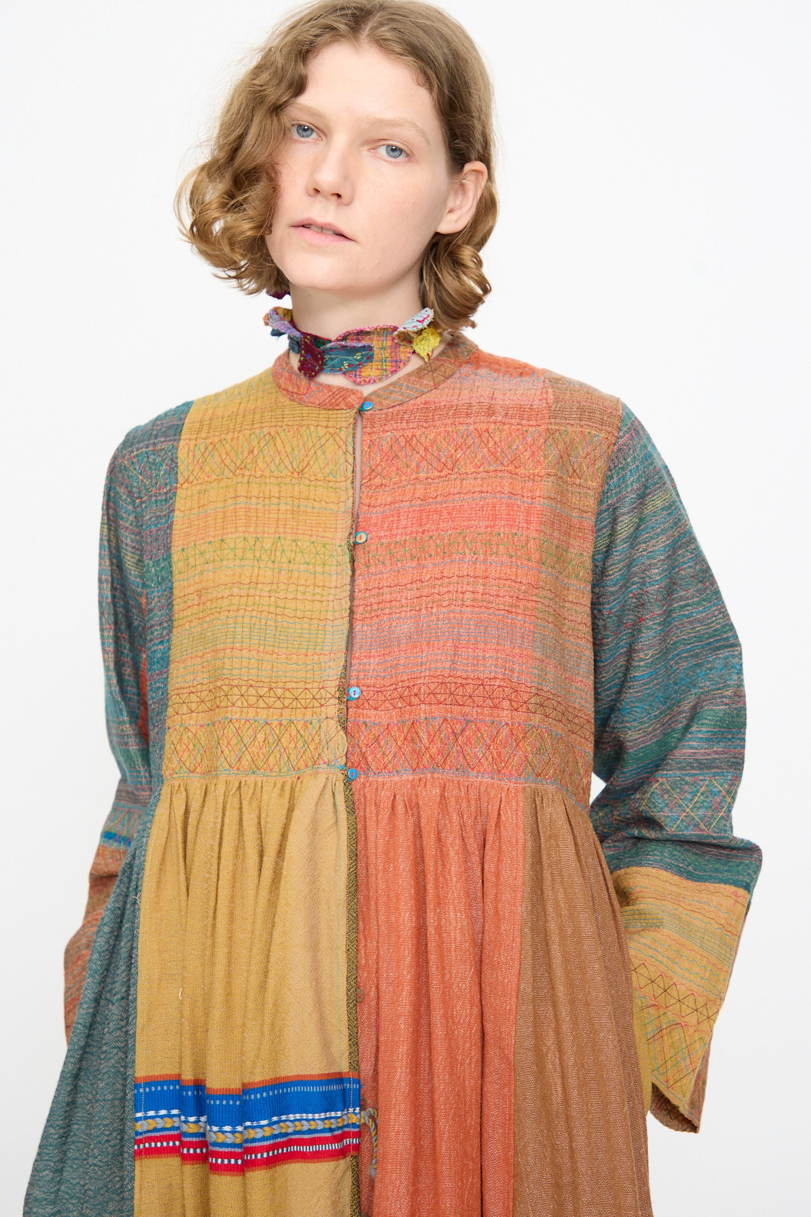 A person with wavy hair wears the Injiri Bhujodi Wool and Silk Boro Jacket in Multi—a long-sleeved, multi-colored patchwork jacket dress featuring a patterned high collar and an intricately embroidered bodice—looking slightly off camera against a plain background.
