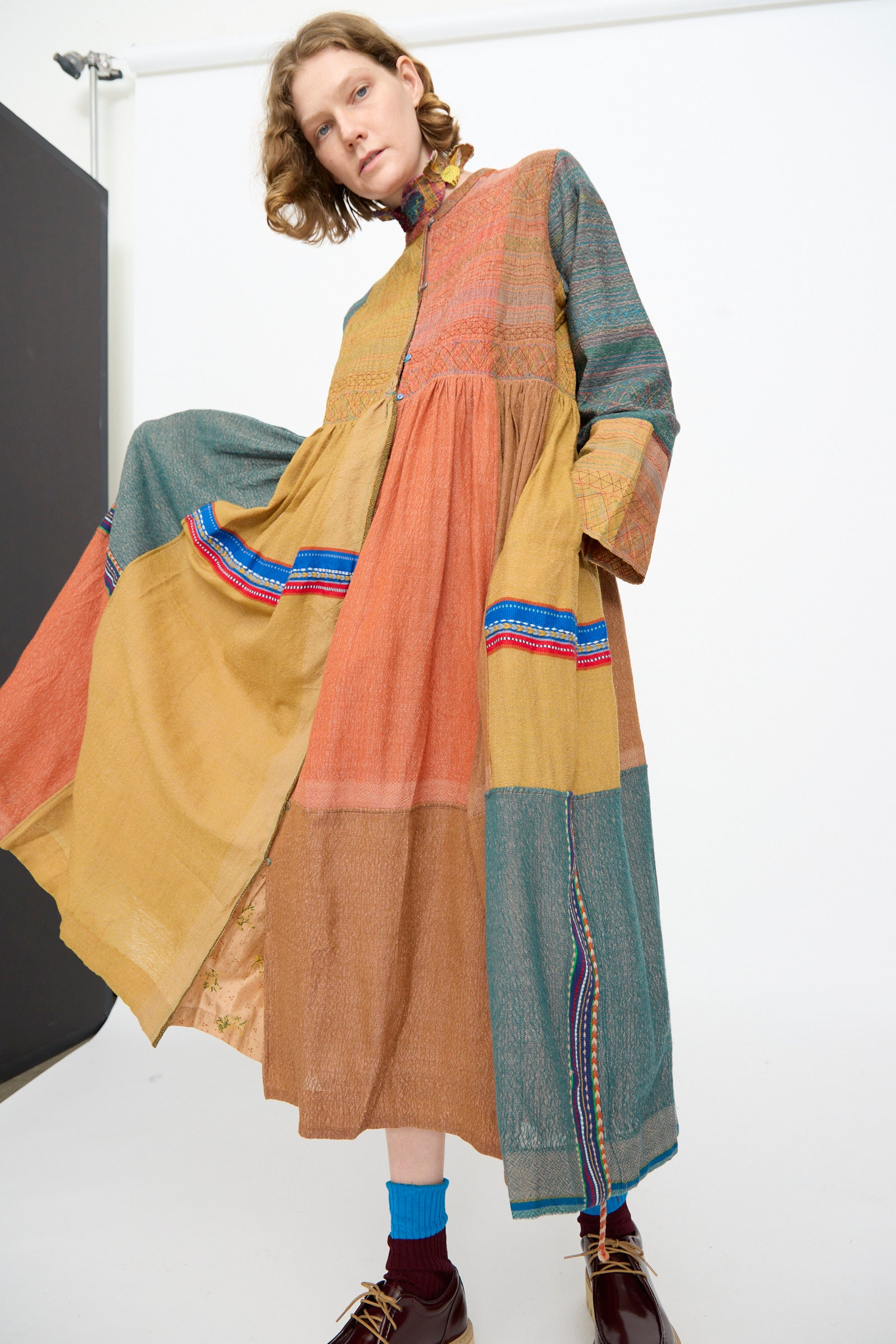 A person wearing the Injiri Bhujodi Wool and Silk Boro Jacket in Multi, paired with blue socks and brown shoes, stands in a studio with a white background.