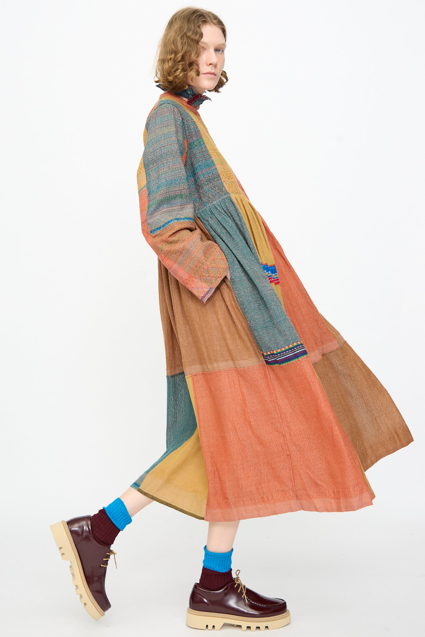 A person wearing the Injiri Bhujodi Wool and Silk Boro Jacket in Multi—featuring a loose, patchwork silhouette in earth tones—paired with an embroidered bodice, blue socks, and brown platform shoes, walks against a plain white background.