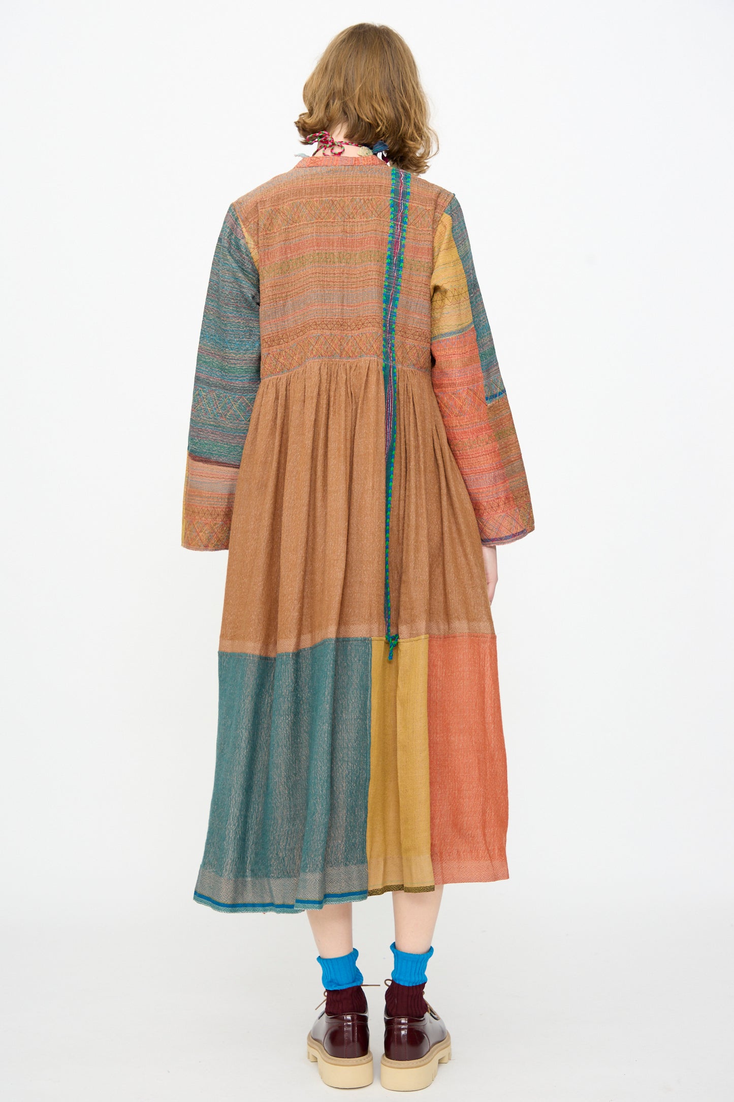 A person stands with their back to the camera, wearing the Injiri Bhujodi Wool and Silk Boro Jacket in Multi—an earth-toned, color-blocked piece featuring textured wool and silk blends. They pair it with teal socks and maroon shoes.