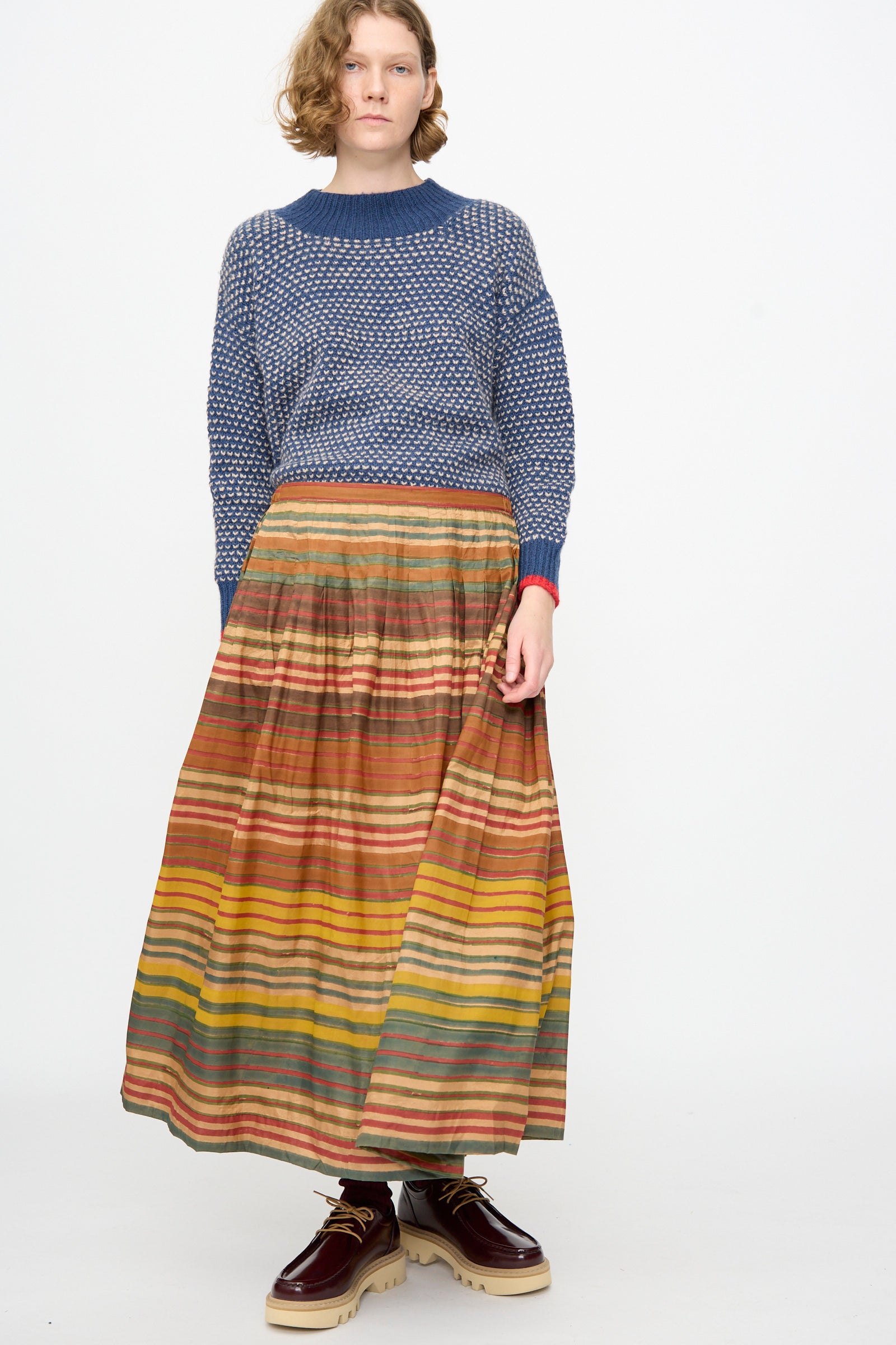 A person stands against a white background, wearing a blue textured sweater, the Injiri Block Print Silk Cotton Zarina Skirt in Stripe Multi, and maroon platform shoes.