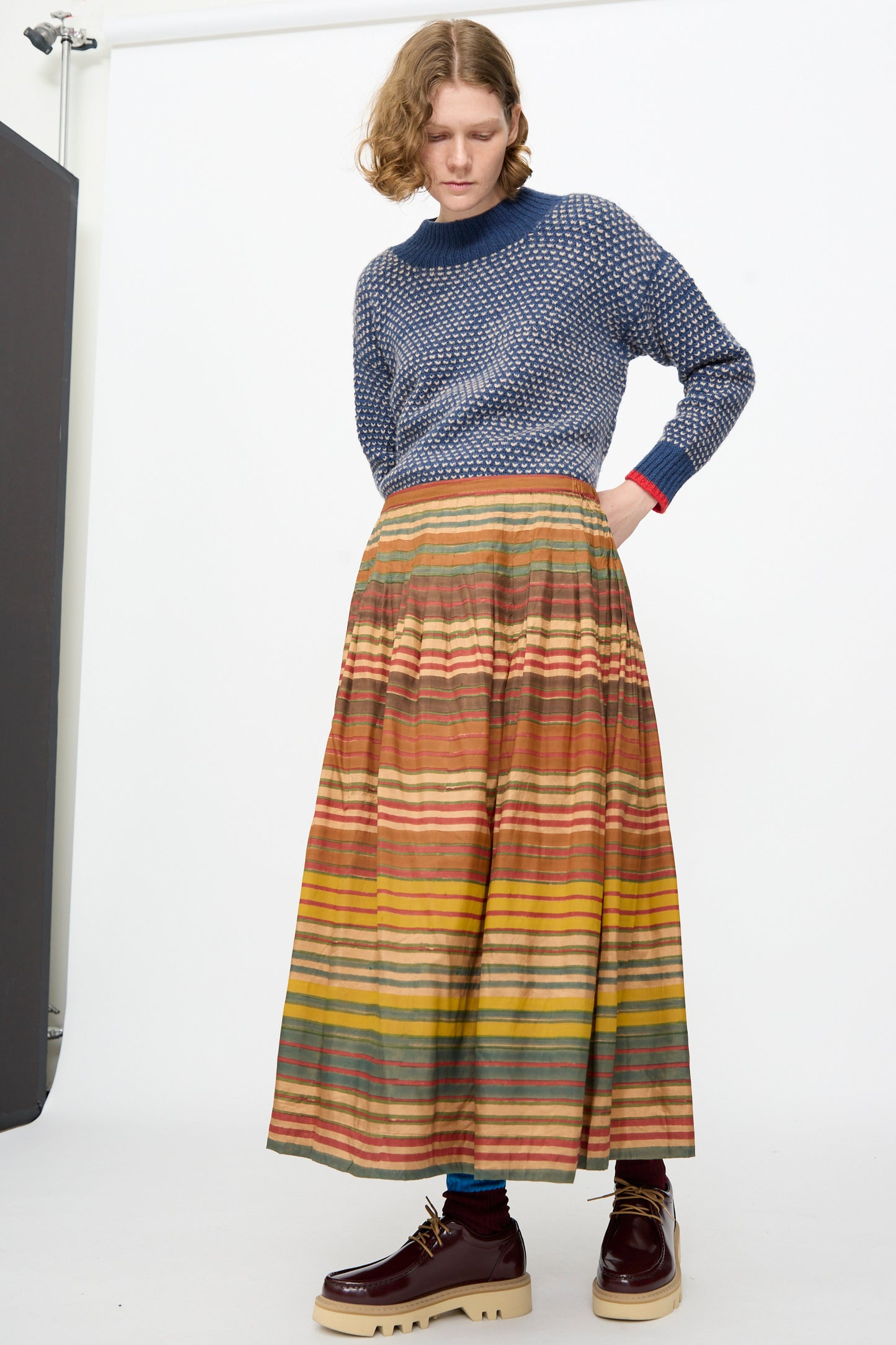A person wearing a navy patterned sweater paired with the Injiri Block Print Silk Cotton Zarina Skirt in Stripe Multi, styled with platform shoes, stands against a plain white backdrop.