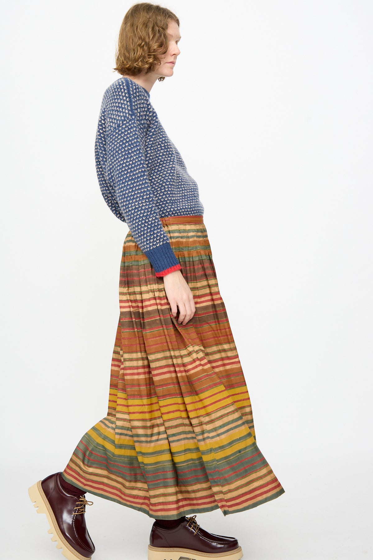 A person in profile wears a blue knit sweater, the Injiri Block Print Silk Cotton Zarina Skirt in Stripe Multi, and maroon platform shoes, walking against a plain white background.