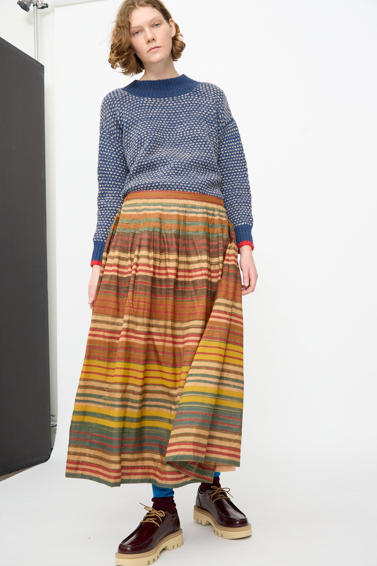 Model wearing a blue textured sweater, the Injiri Block Print Silk Cotton Zarina Skirt in Stripe Multi, blue socks, and brown platform shoes, standing against a plain white background.