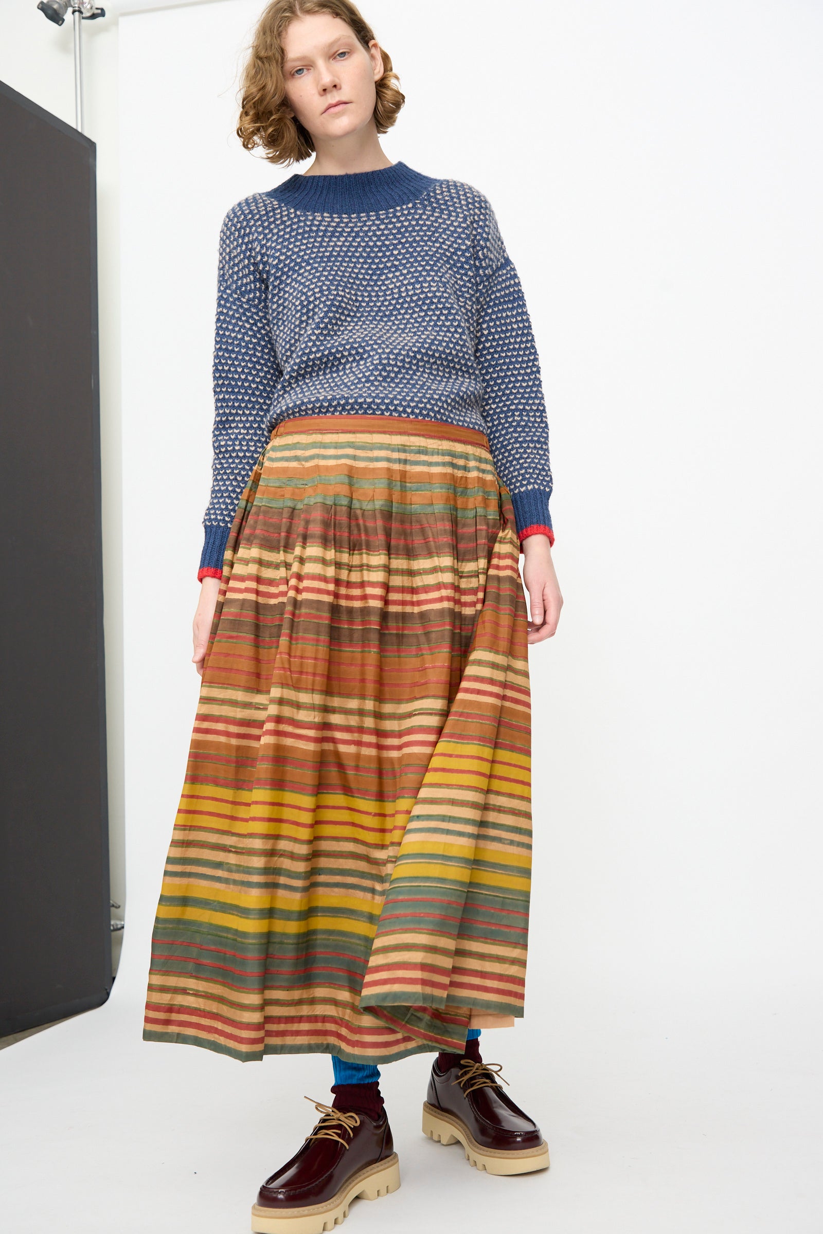 Model wearing a blue textured sweater, the Injiri Block Print Silk Cotton Zarina Skirt in Stripe Multi, blue socks, and brown platform shoes, standing against a plain white background.