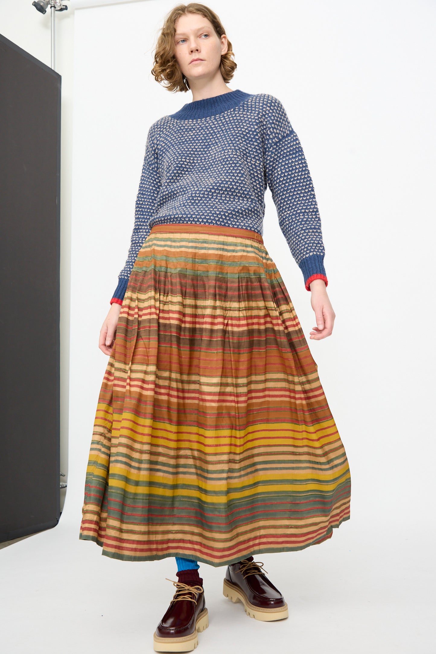 A person stands against a plain white background wearing a blue textured sweater, blue socks, brown shoes, and the Injiri Block Print Silk Cotton Zarina Skirt in Stripe Multi.
