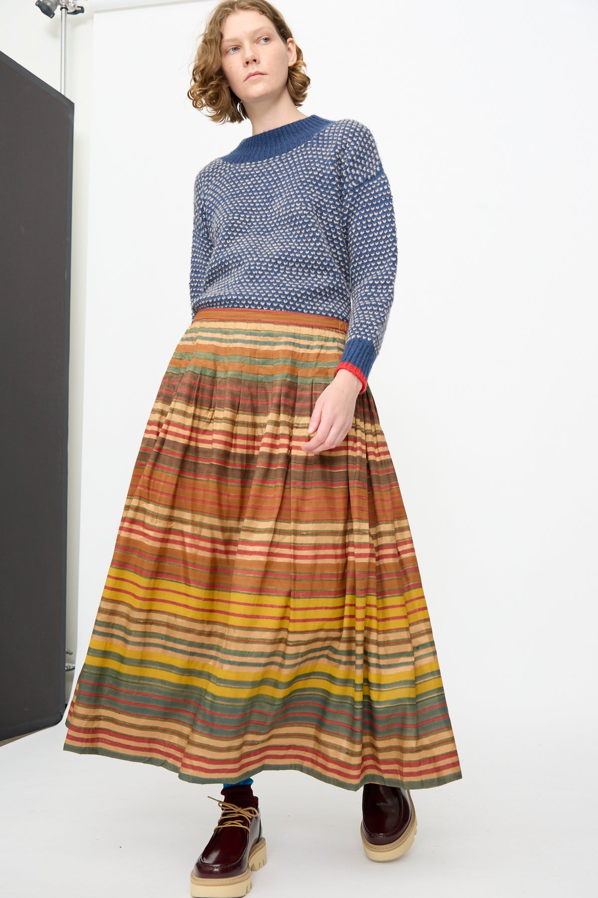 A person stands against a white wall wearing a blue textured sweater, blue tights, brown platform shoes, and the Injiri Block Print Silk Cotton Zarina Skirt in Stripe Multi.
