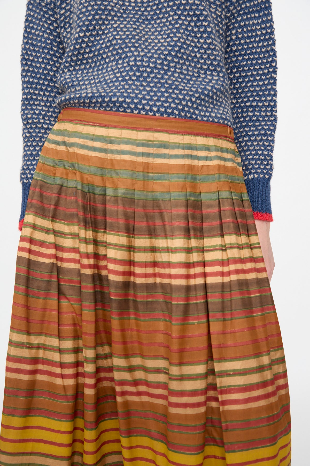 Person wearing a blue patterned sweater and the Injiri Block Print Silk Cotton Zarina Skirt in Stripe Multi, featuring pleated horizontal stripes in brown, red, yellow, and green tones.