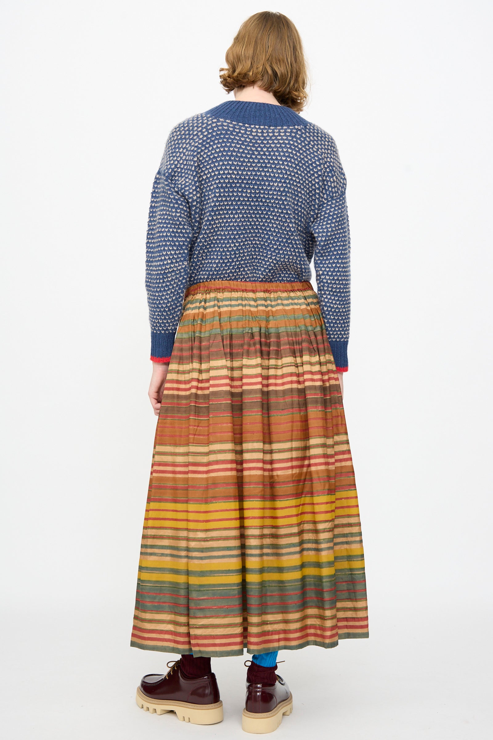 A person stands with their back to the camera, dressed in a patterned blue sweater and the Injiri Block Print Silk Cotton Zarina Skirt in Stripe Multi, paired with teal socks and brown platform shoes against a plain white background.