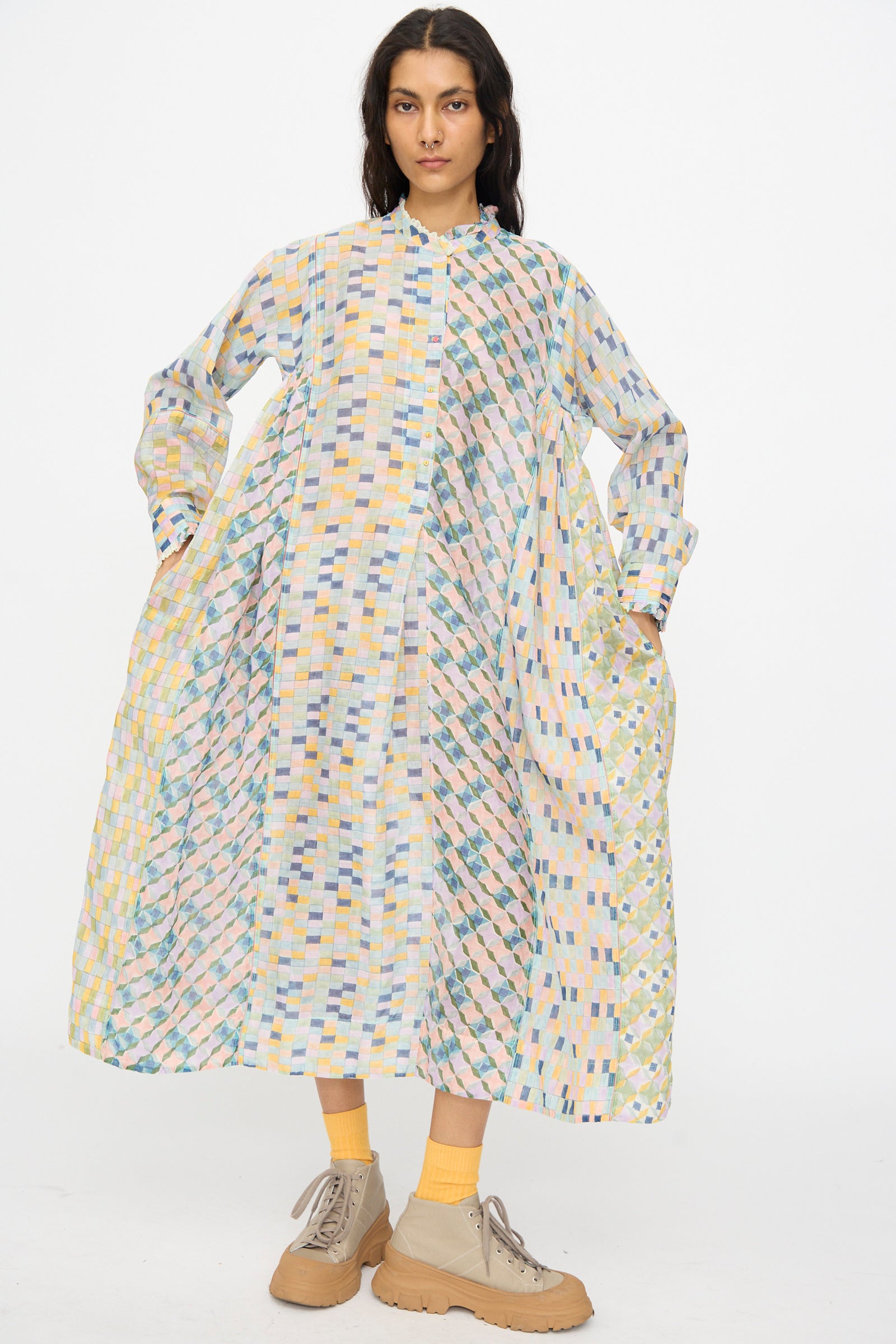 A woman stands with hands in her pockets, wearing the Injiri Blockprint Chanderi Trumpet Dress in Blue Yellow Multi, paired with yellow socks and beige shoes against a plain white background.