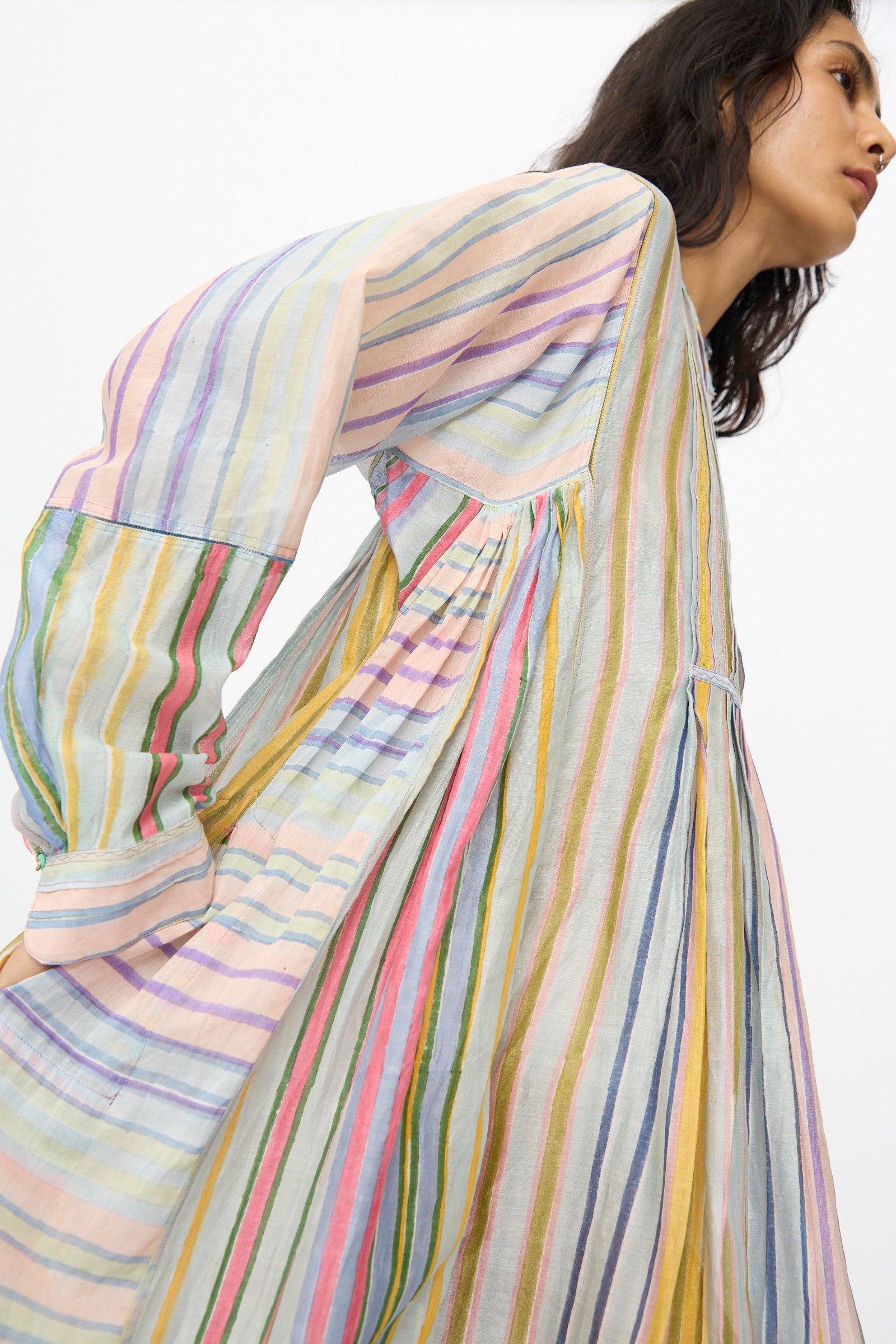A woman wears the Injiri Blockprint Chanderi Trumpet Dress in Multi Stripe, a colorful midi dress with loose, long sleeves, shown from the side against a plain background.