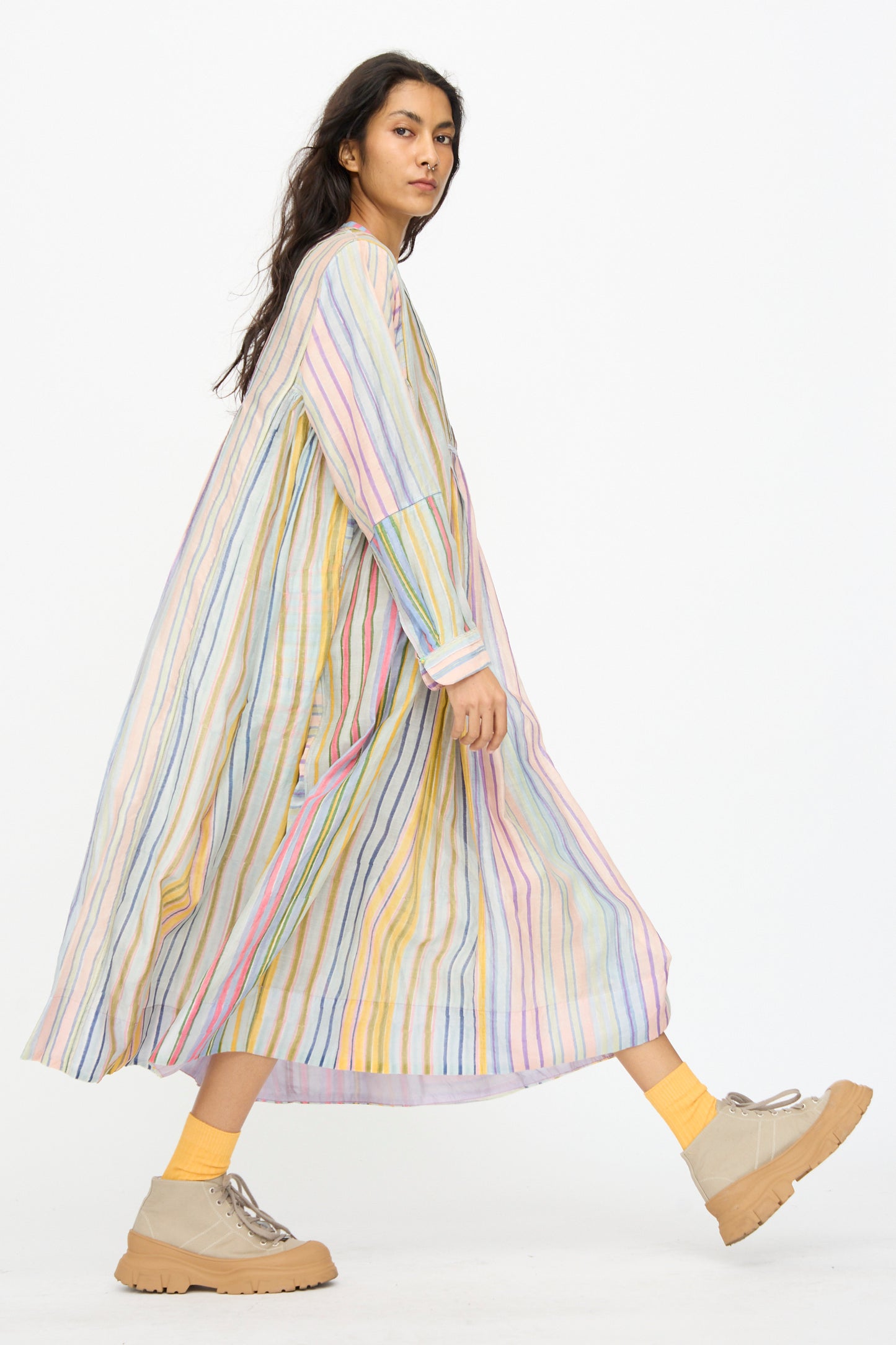 A person walks against a plain white background wearing the Injiri Blockprint Chanderi Trumpet Dress in Multi Stripe, styled with yellow socks and beige shoes.