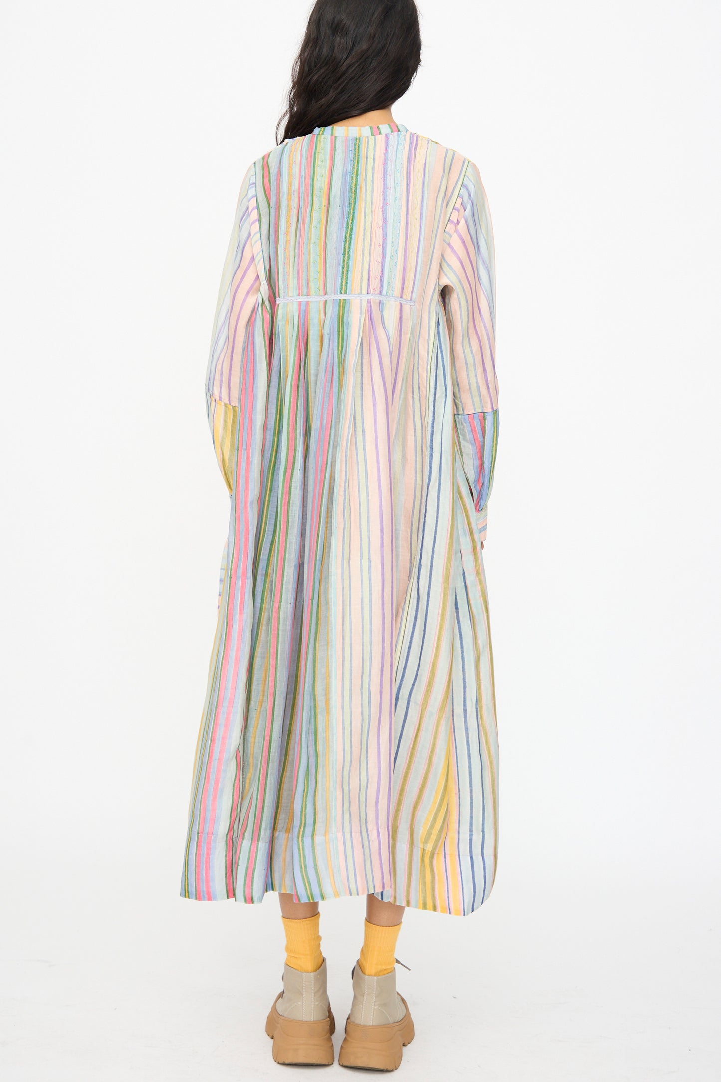 A person faces away, wearing the Injiri Blockprint Chanderi Trumpet Dress in Multi Stripe—an ankle-length, long-sleeved dress—paired with yellow socks and beige shoes, set against a white background.