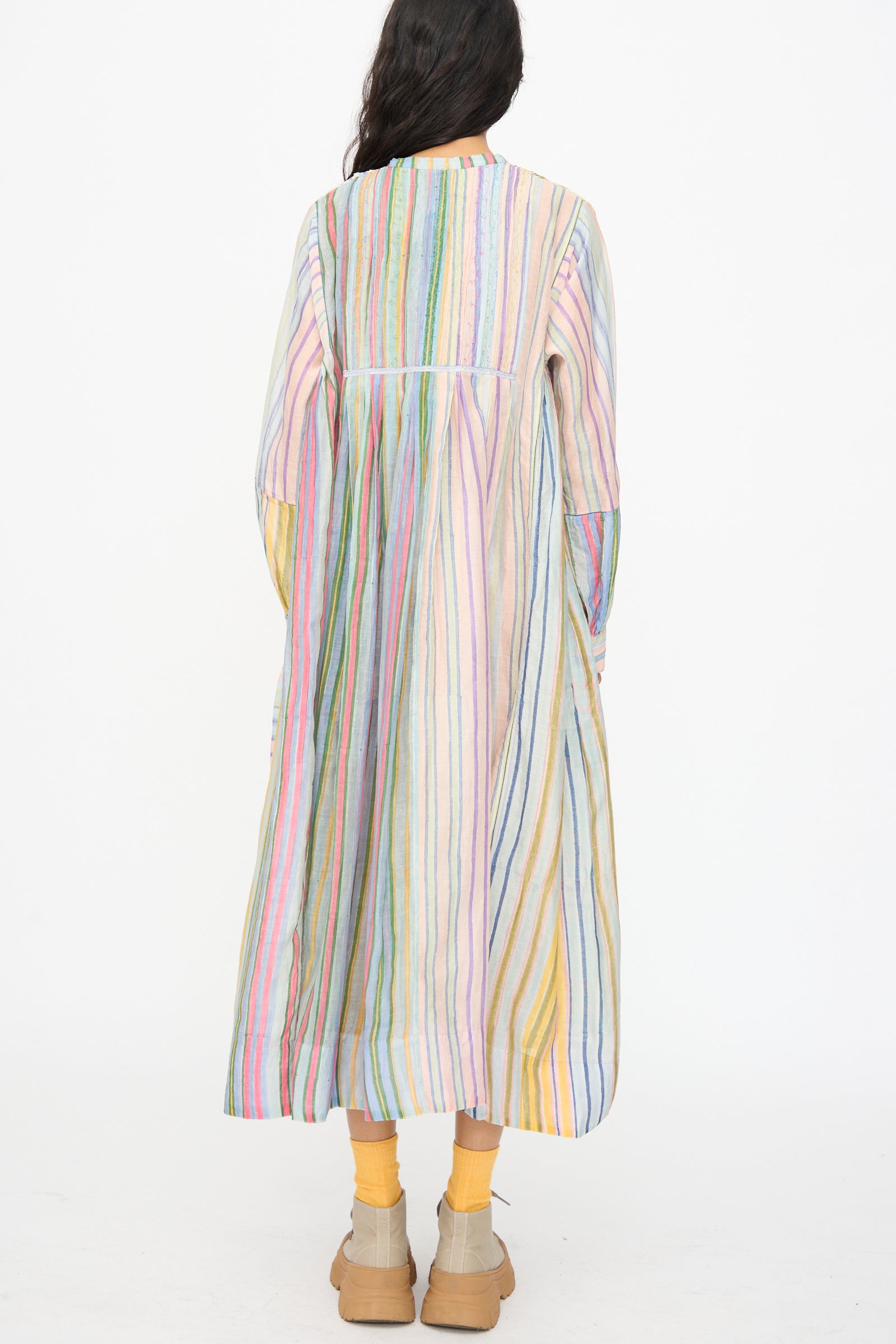 A person faces away, wearing the Injiri Blockprint Chanderi Trumpet Dress in Multi Stripe—an ankle-length, long-sleeved dress—paired with yellow socks and beige shoes, set against a white background.