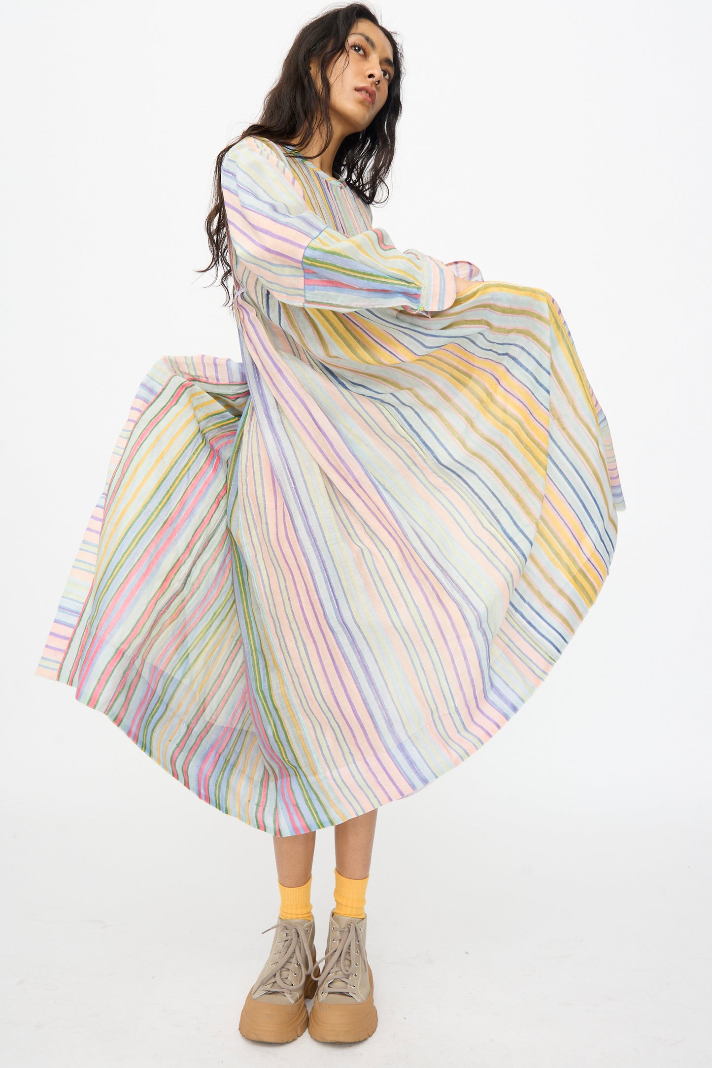 A model in Injiri’s Blockprint Chanderi Trumpet Dress in Multi Stripe stands against a white background, holding out the skirt, paired with yellow socks and beige shoes.
