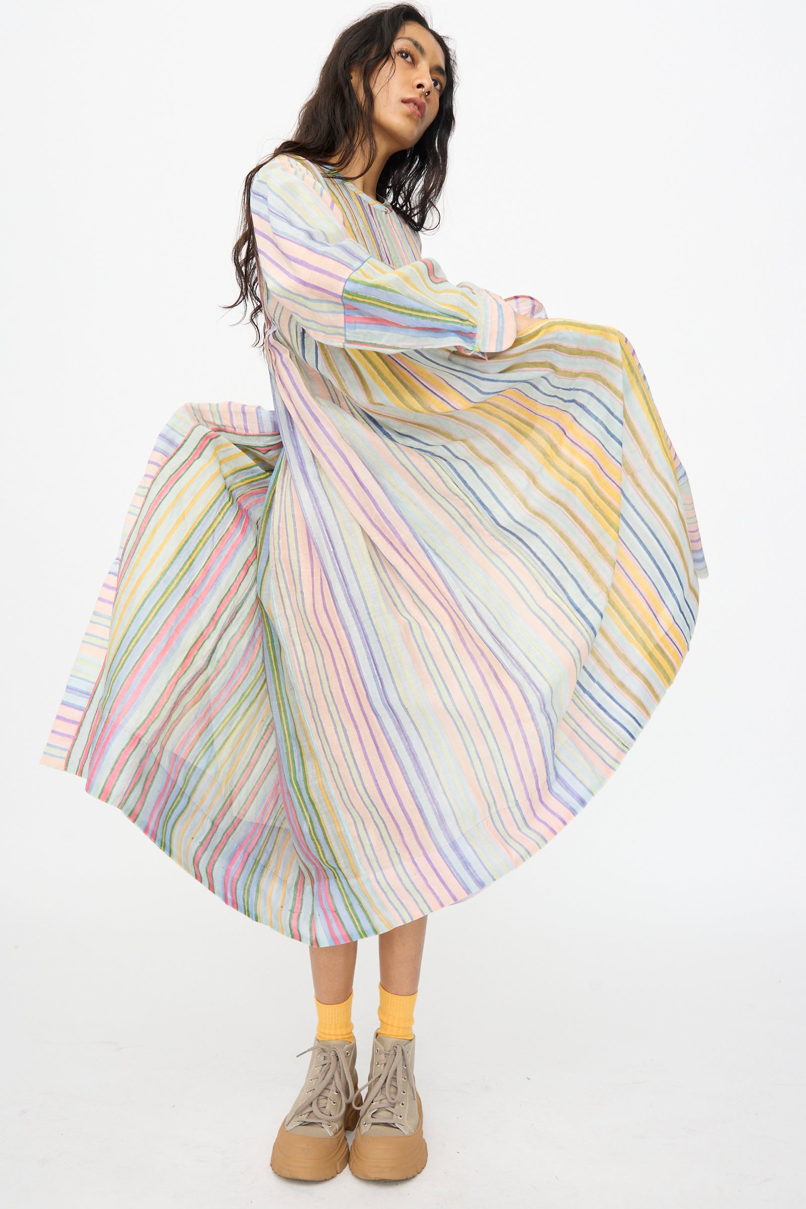 A model in Injiri’s Blockprint Chanderi Trumpet Dress in Multi Stripe stands against a white background, holding out the skirt, paired with yellow socks and beige shoes.