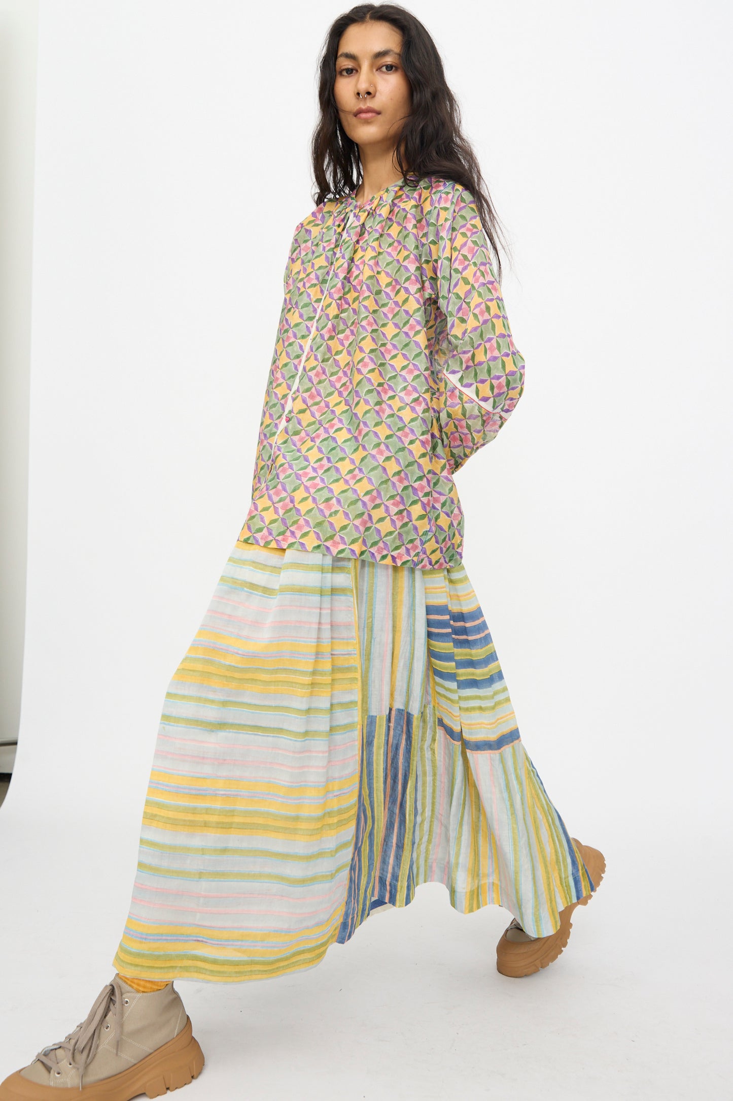 A person in the Injiri Blockprint Chanderi Zamila Skirt in Green Multi, paired with a patterned long-sleeve top and tan sneakers, stands against a plain white background.