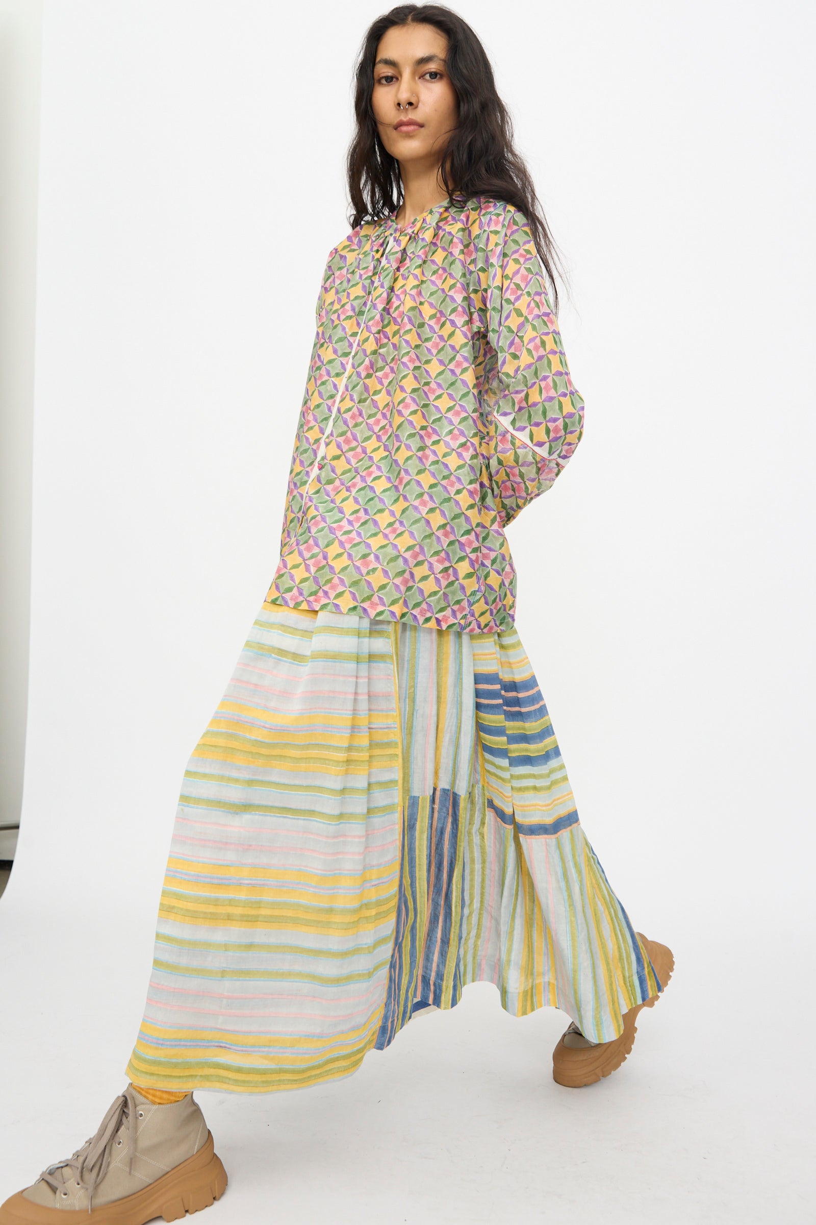 A person in the Injiri Blockprint Chanderi Zamila Skirt in Green Multi, paired with a patterned long-sleeve top and tan sneakers, stands against a plain white background.