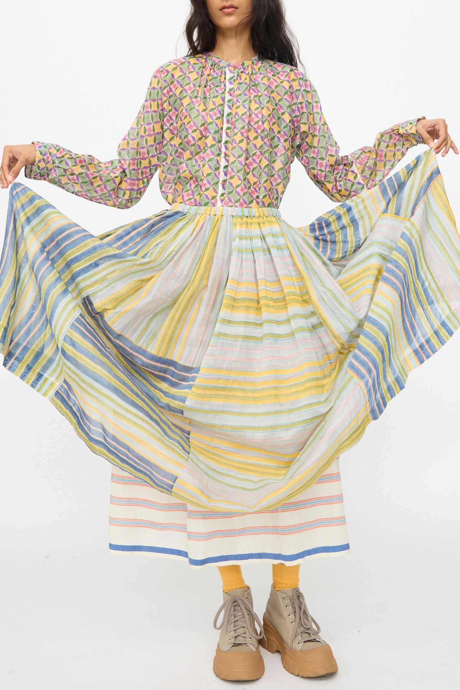 A person wears a patterned long-sleeve blouse, yellow socks, tan platform shoes, and holds out the Injiri Blockprint Chanderi Zamila Skirt in Green Multi.