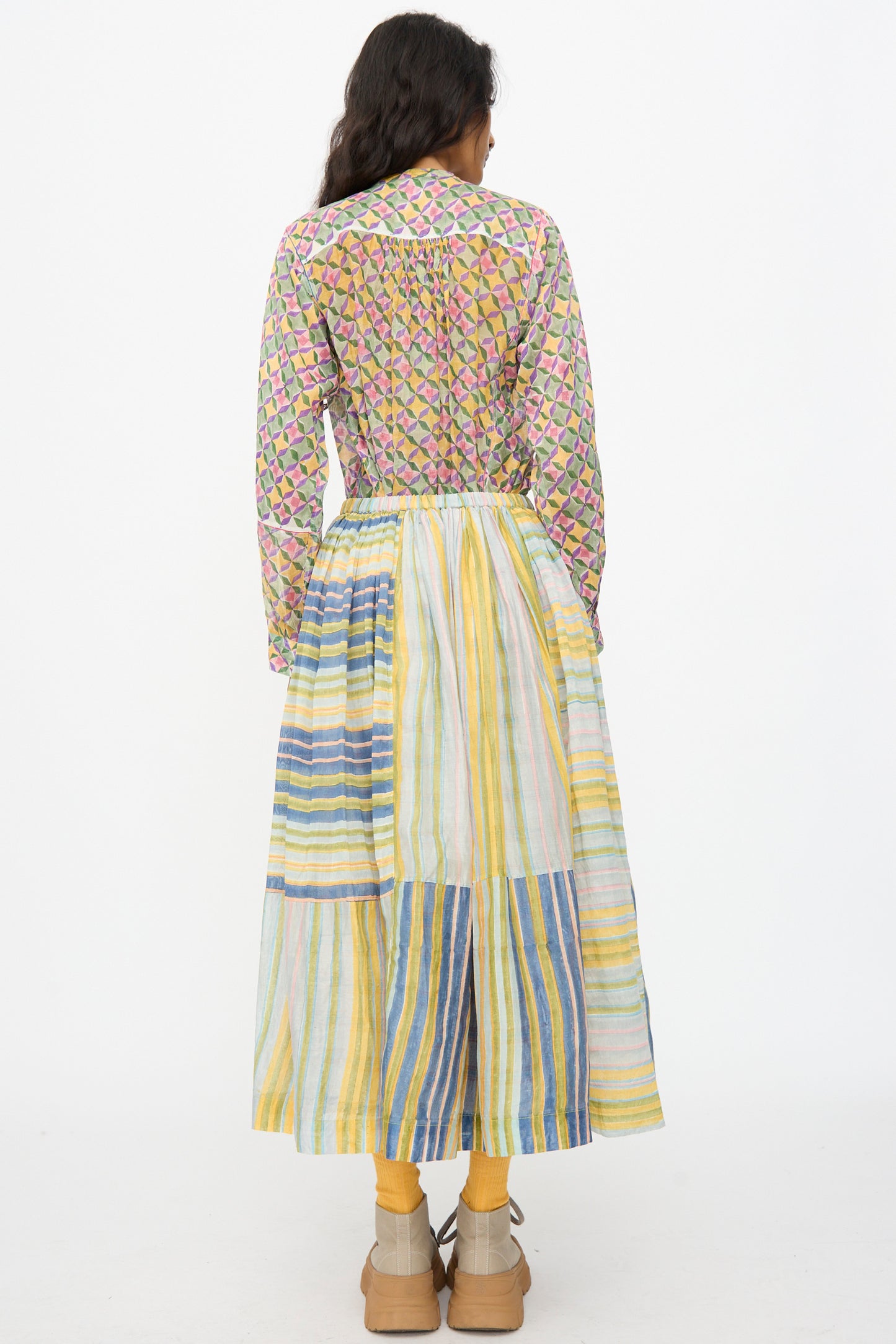 A person faces away, wearing the Injiri Blockprint Chanderi Zamila Skirt in Green Multi with a patterned long-sleeve shirt, yellow socks, and tan shoes against a plain white background.