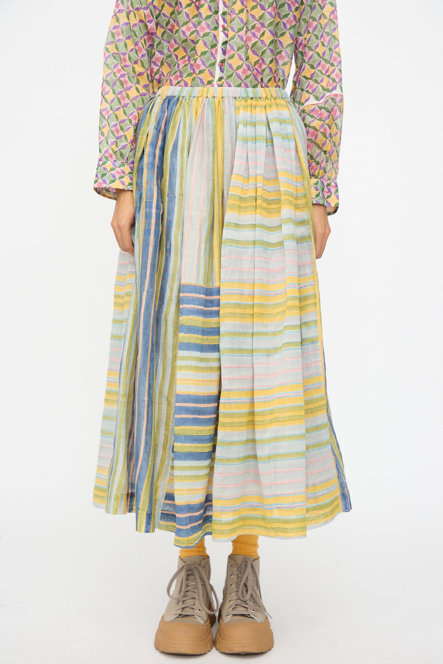 A person wears the Injiri Blockprint Chanderi Zamila Skirt in Green Multi with a patterned long-sleeve top and tan lace-up shoes, standing against a plain white background.