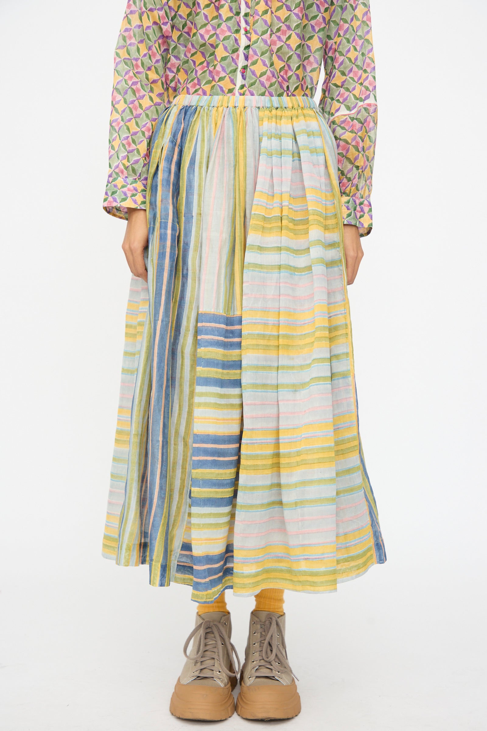 A person wears the Injiri Blockprint Chanderi Zamila Skirt in Green Multi with a patterned long-sleeve top and tan lace-up shoes, standing against a plain white background.