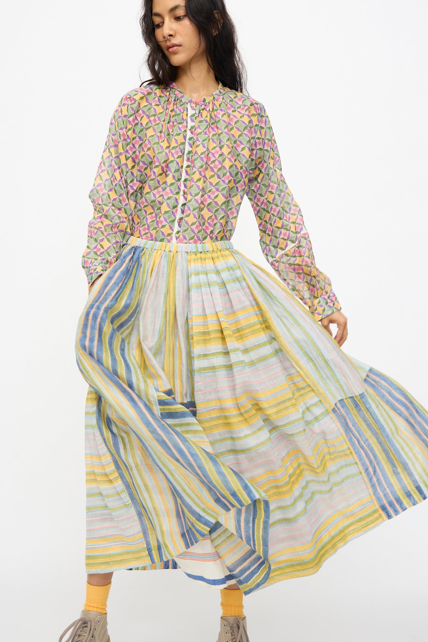 A woman models the Injiri Blockprint Chanderi Zamila Skirt in Green Multi, featuring a vibrant patchwork design, standing against a plain white background and holding the skirt out to the side.