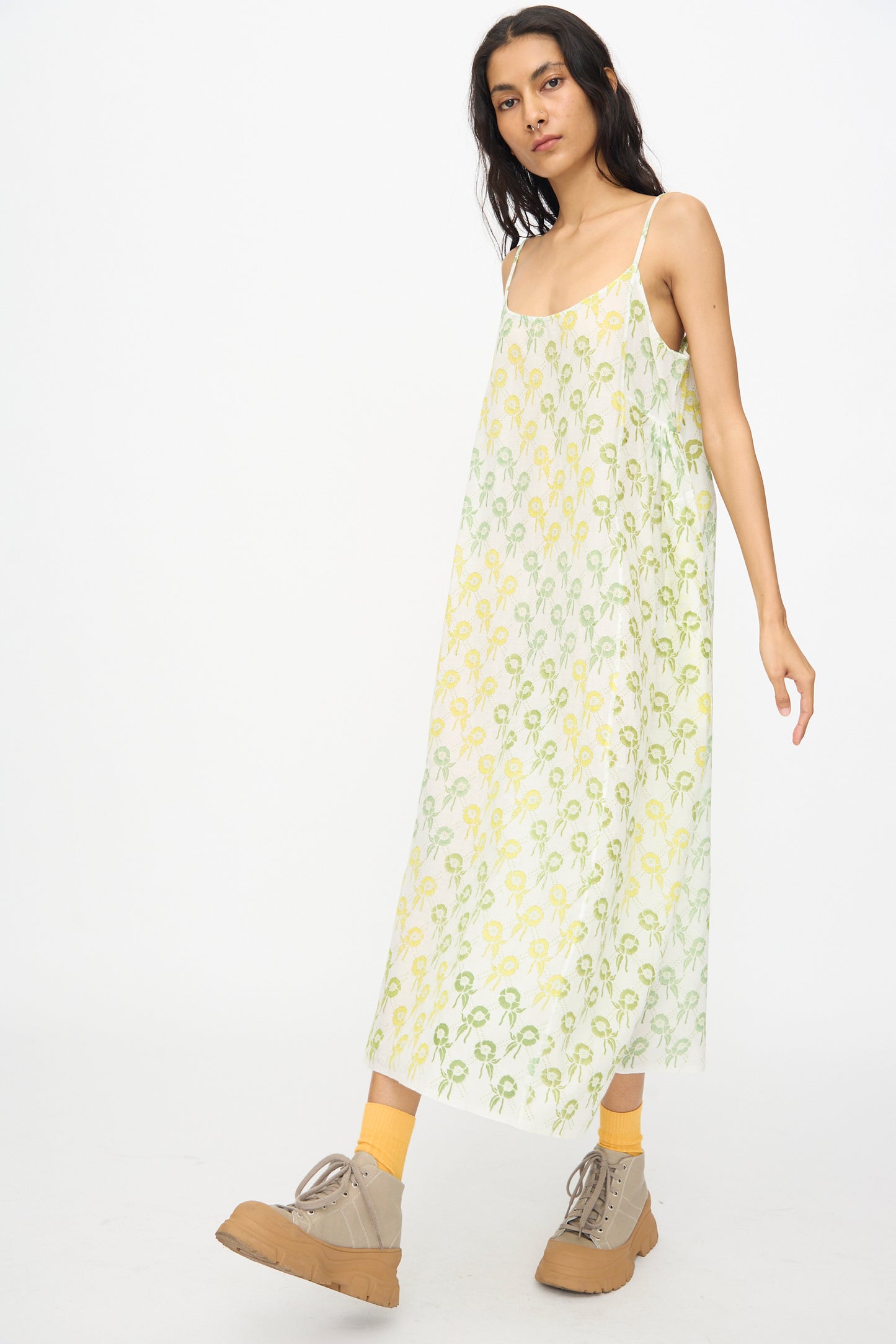 A woman wears the Injiri Blockprint Cotton Emma Slip in Green Multi— a loose, block-printed cotton midi dress—paired with yellow socks and beige lace-up shoes, set against a plain white background.