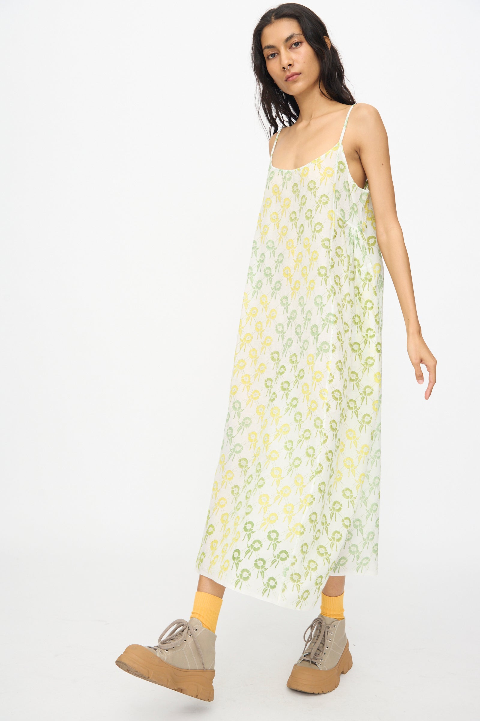 A woman wears the Injiri Blockprint Cotton Emma Slip in Green Multi— a loose, block-printed cotton midi dress—paired with yellow socks and beige lace-up shoes, set against a plain white background.