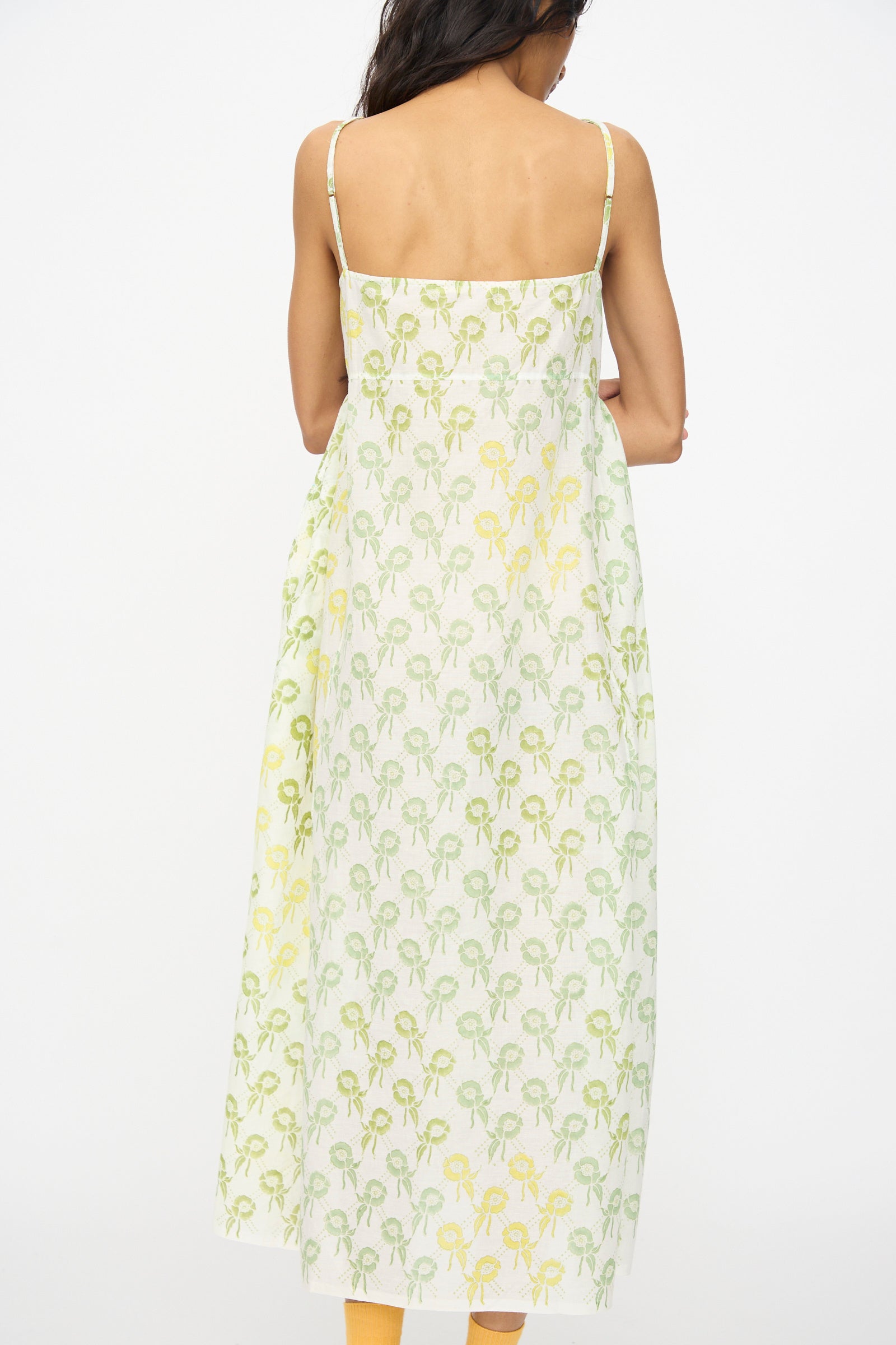 A woman faces away, wearing the Injiri Blockprint Cotton Emma Slip in Green Multi—a sleeveless, thin-strapped midi dress featuring a green and yellow floral block print on white cotton.