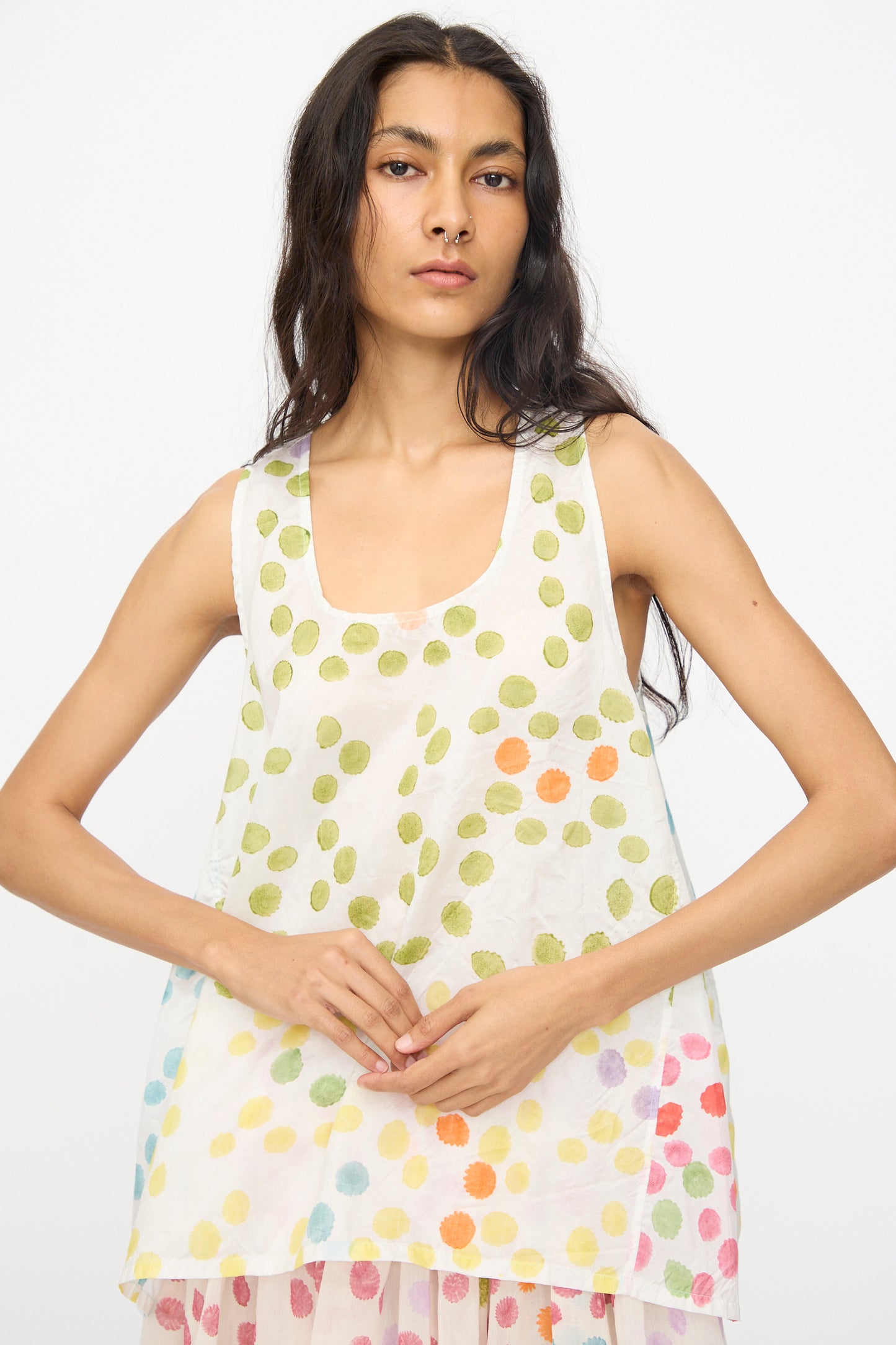 A woman wearing the Injiri Blockprint Silk Bandi Top in White Multi, a loose sleeveless silk piece with a colorful dotted pattern, stands with her hands clasped, facing forward against a plain background.