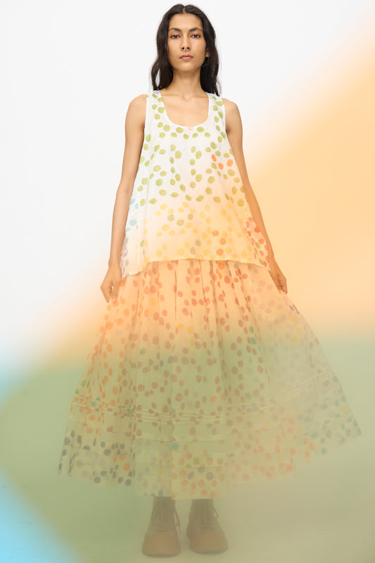 A woman faces forward, wearing a white sleeveless top and the Injiri Blockprint Silk Cotton Zamila Skirt in White Dot Multi. The foreground features an orange and blue blur effect.