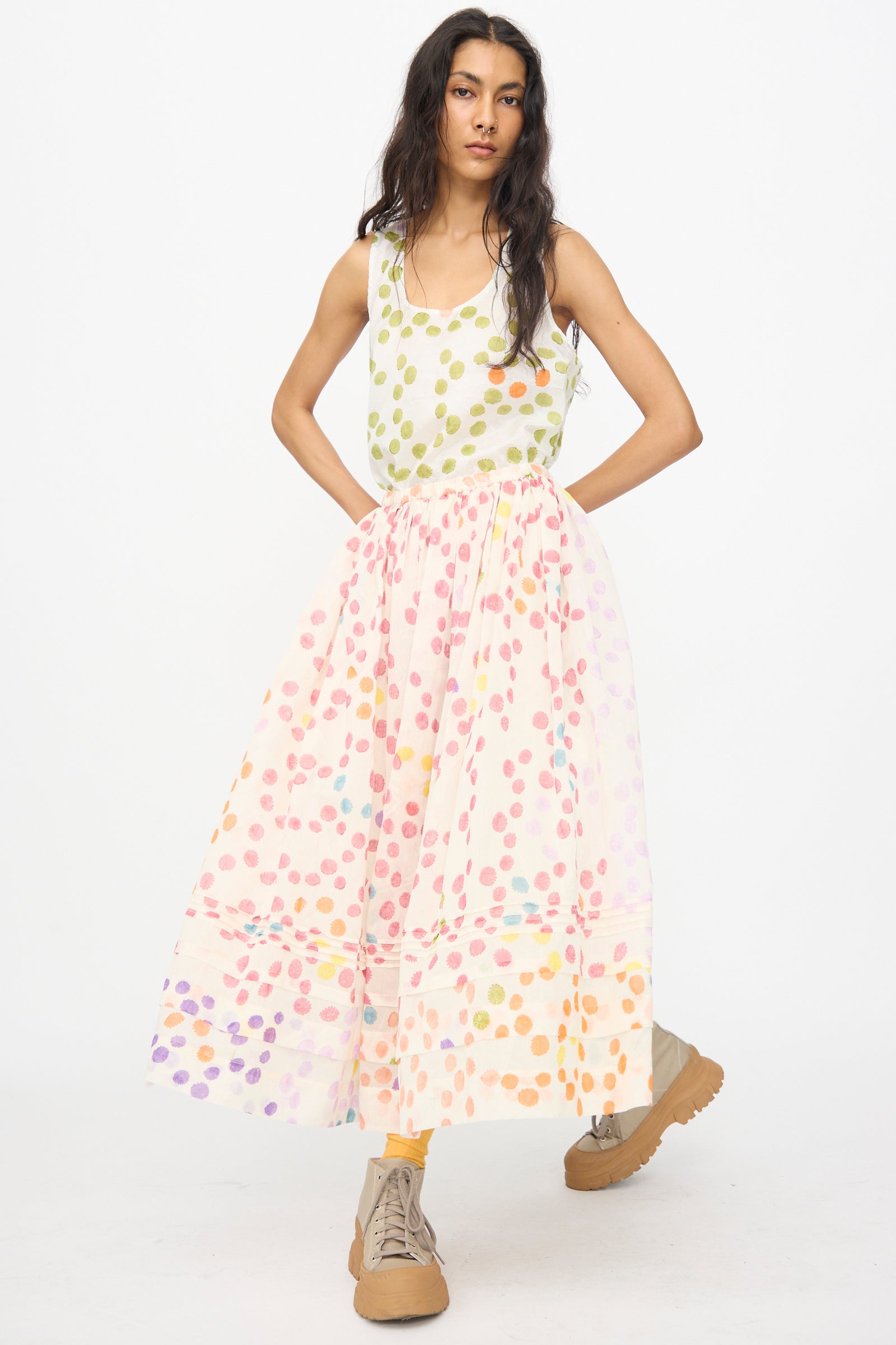 A person stands against a plain background wearing a sleeveless white top with green polka dots, the Injiri Blockprint Silk Cotton Zamila Skirt in White Dot Multi, tan boots, and yellow socks.