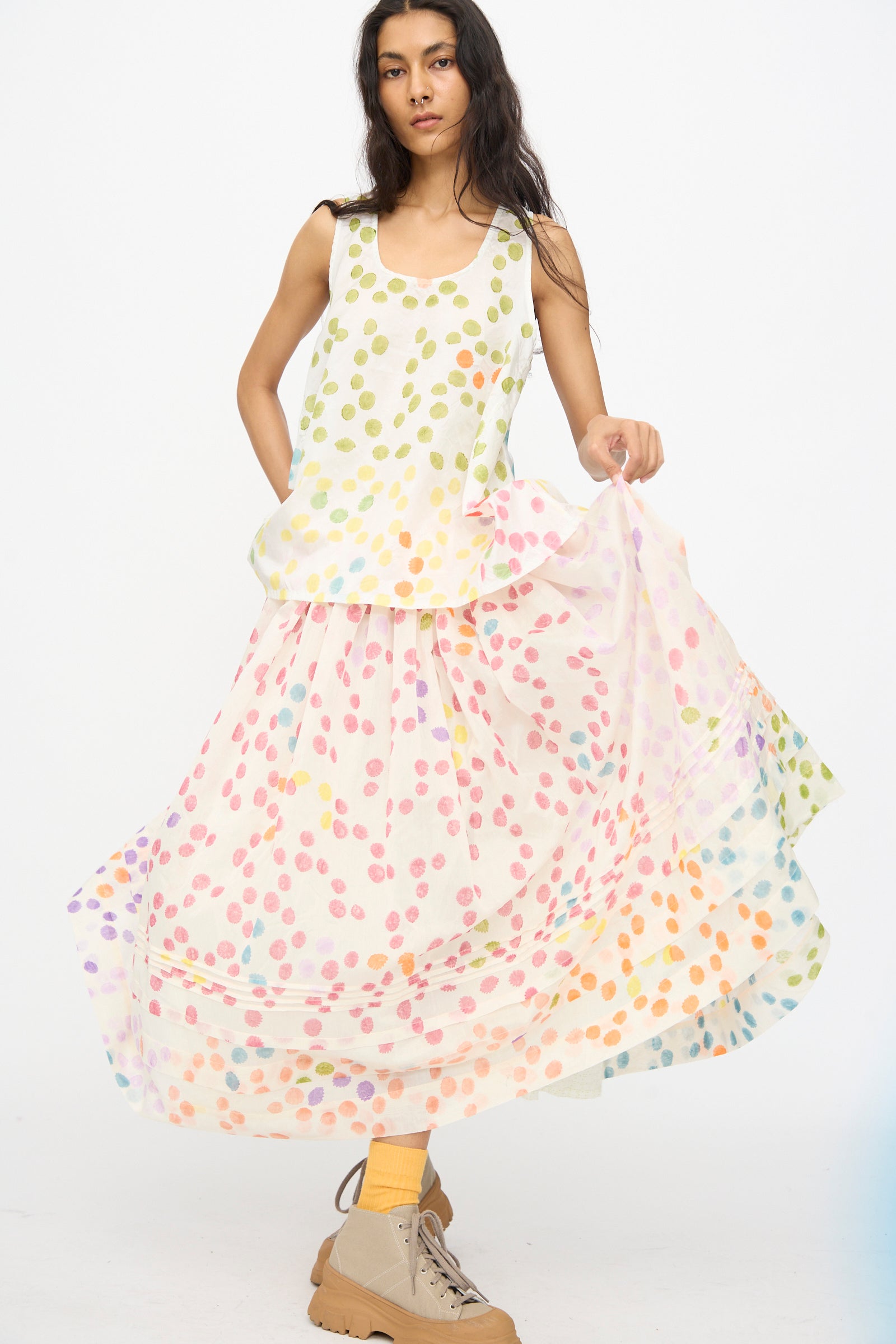 A woman wears the Injiri Blockprint Silk Cotton Zamila Skirt in White Dot Multi, styled with a sleeveless top, tan boots, and yellow socks, standing against a plain white background.