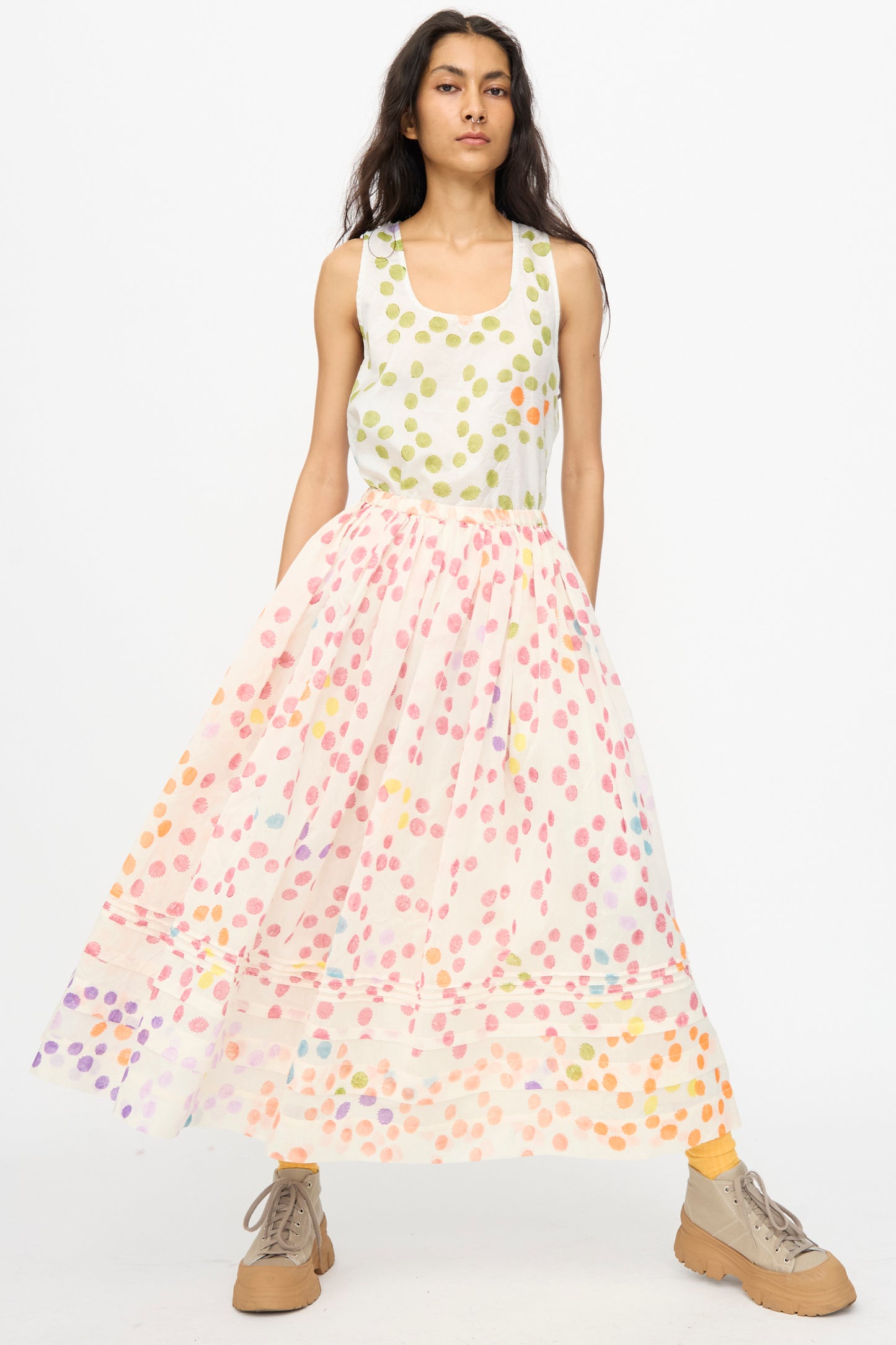 A woman wears the Injiri Blockprint Silk Cotton Zamila Skirt in White Dot Multi, a flowing midi skirt with multicolor polka dots, paired with a sleeveless white top and tan boots.