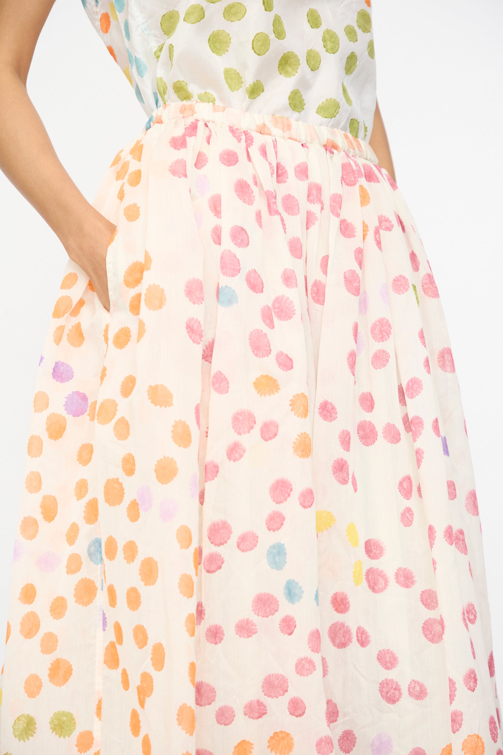 A person models the Injiri Blockprint Silk Cotton Zamila Skirt in White Dot Multi, featuring multicolored dots, shown from shoulders to knees with one hand in a pocket against a plain background.