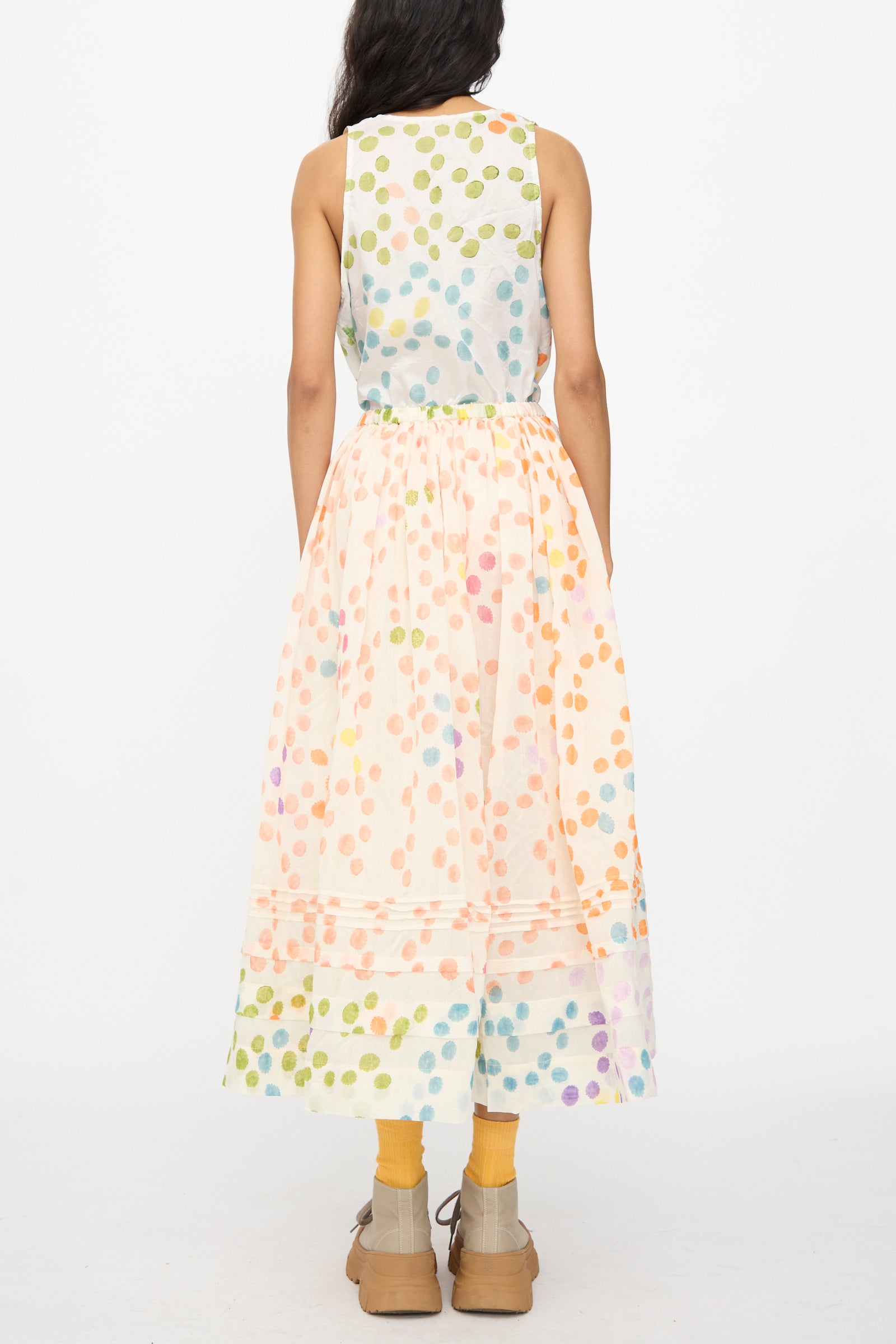 A person faces away, wearing a sleeveless top and the Injiri Blockprint Silk Cotton Zamila Skirt in White Dot Multi, yellow socks, and beige platform shoes against a plain white background.