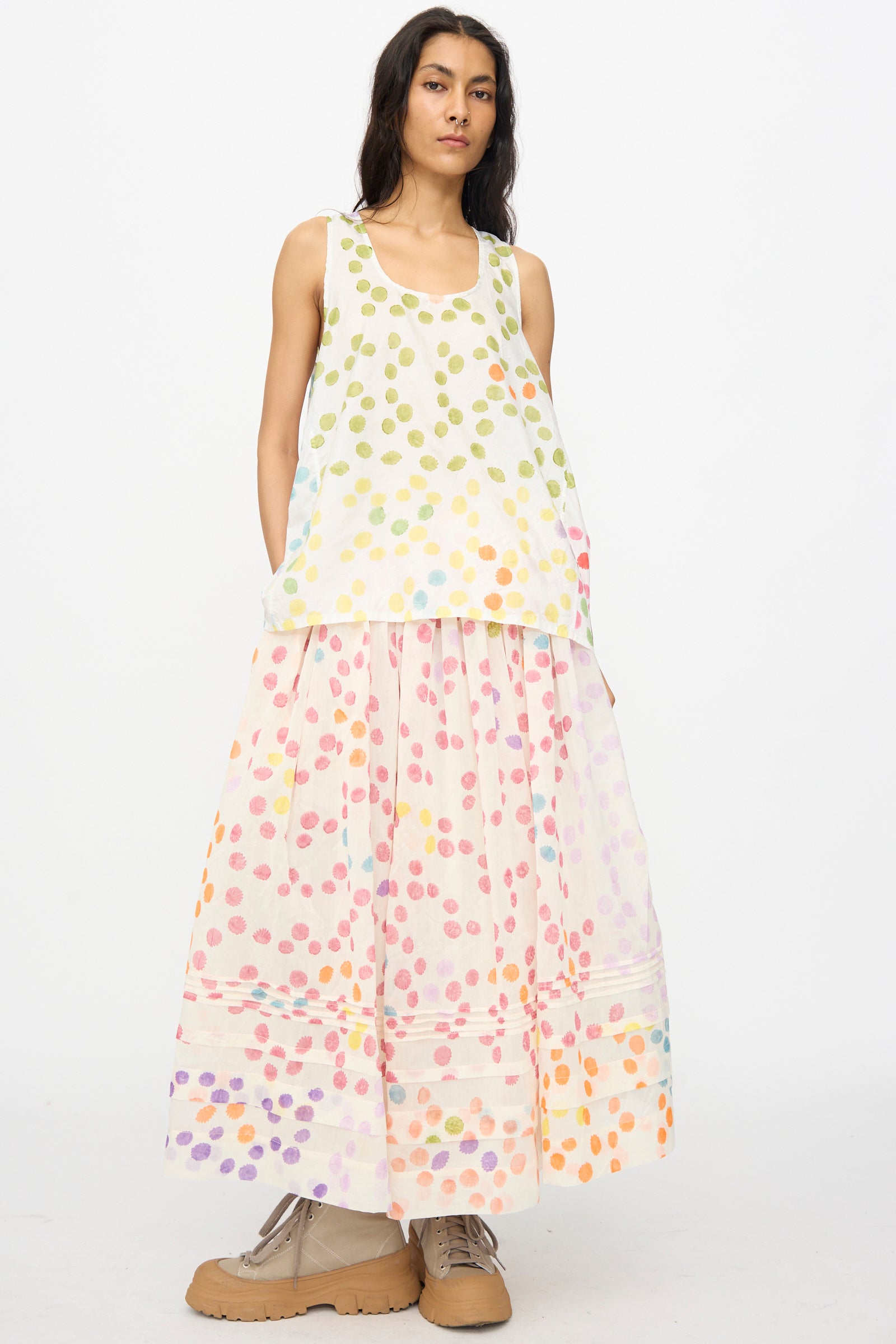 A woman wears the Injiri Blockprint Silk Cotton Zamila Skirt in White Dot Multi—a long, full, white skirt with multicolored polka dots—paired with a sleeveless top and tan platform shoes against a plain background.