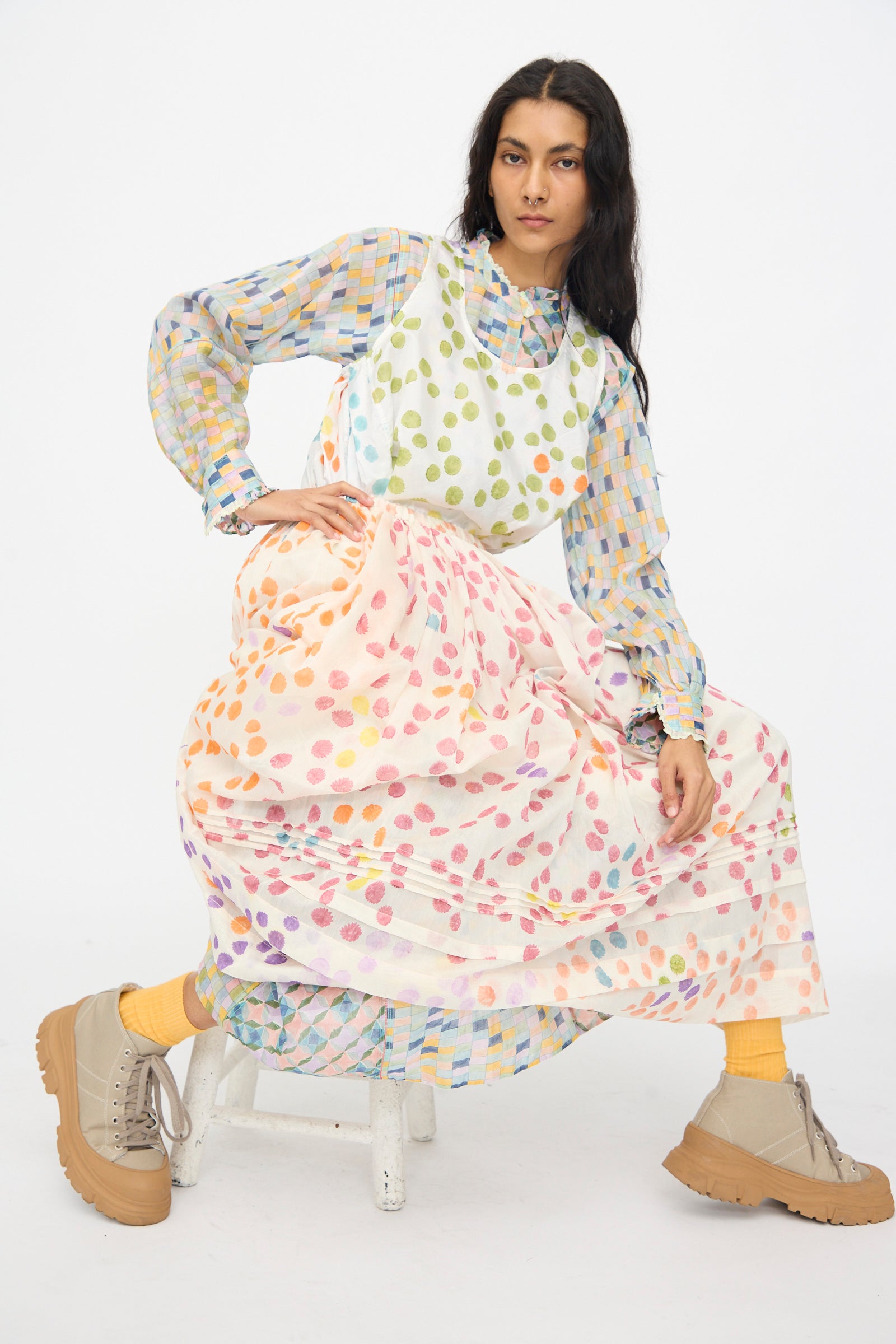 Woman wearing a Injiri's Blockprint Chanderi Trumpet Dress in Blue Yellow Multi, with the Blockprint Silk Bandi Top in White Multi over it, and the Blockprint Silk Cotton Zamila Skirt in White Dot Multi sitting on a stool against a white background.