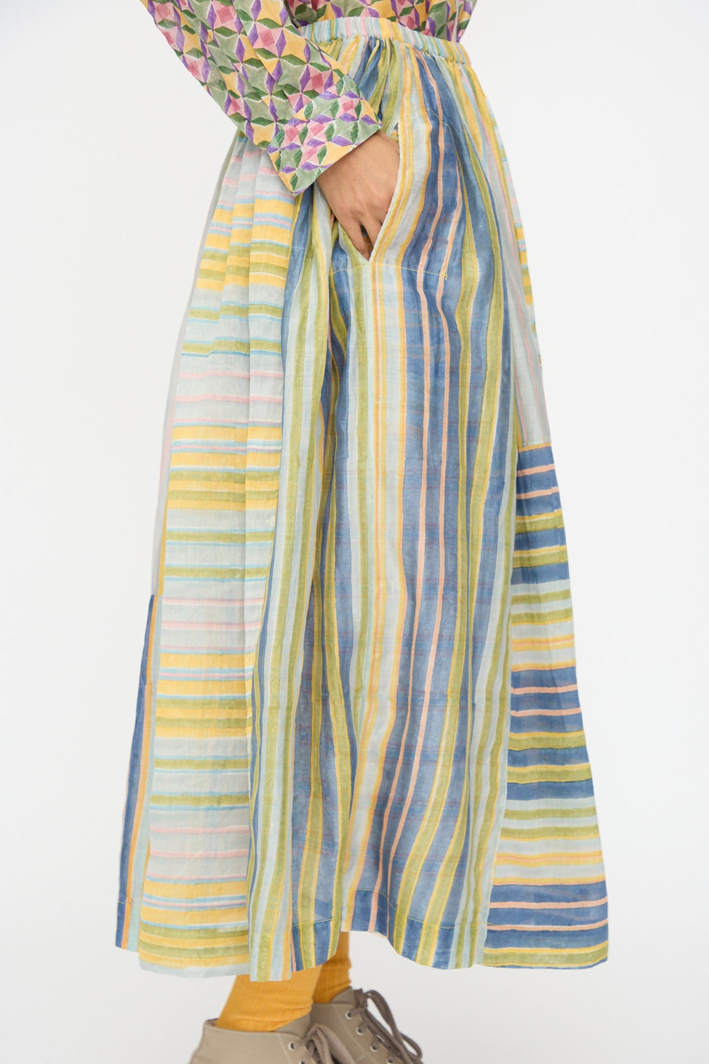 A person wears the Injiri Blockprint Chanderi Zamila Skirt in Green Multi with a hand in the pocket, paired with yellow tights, beige shoes, and a patterned long-sleeve top.