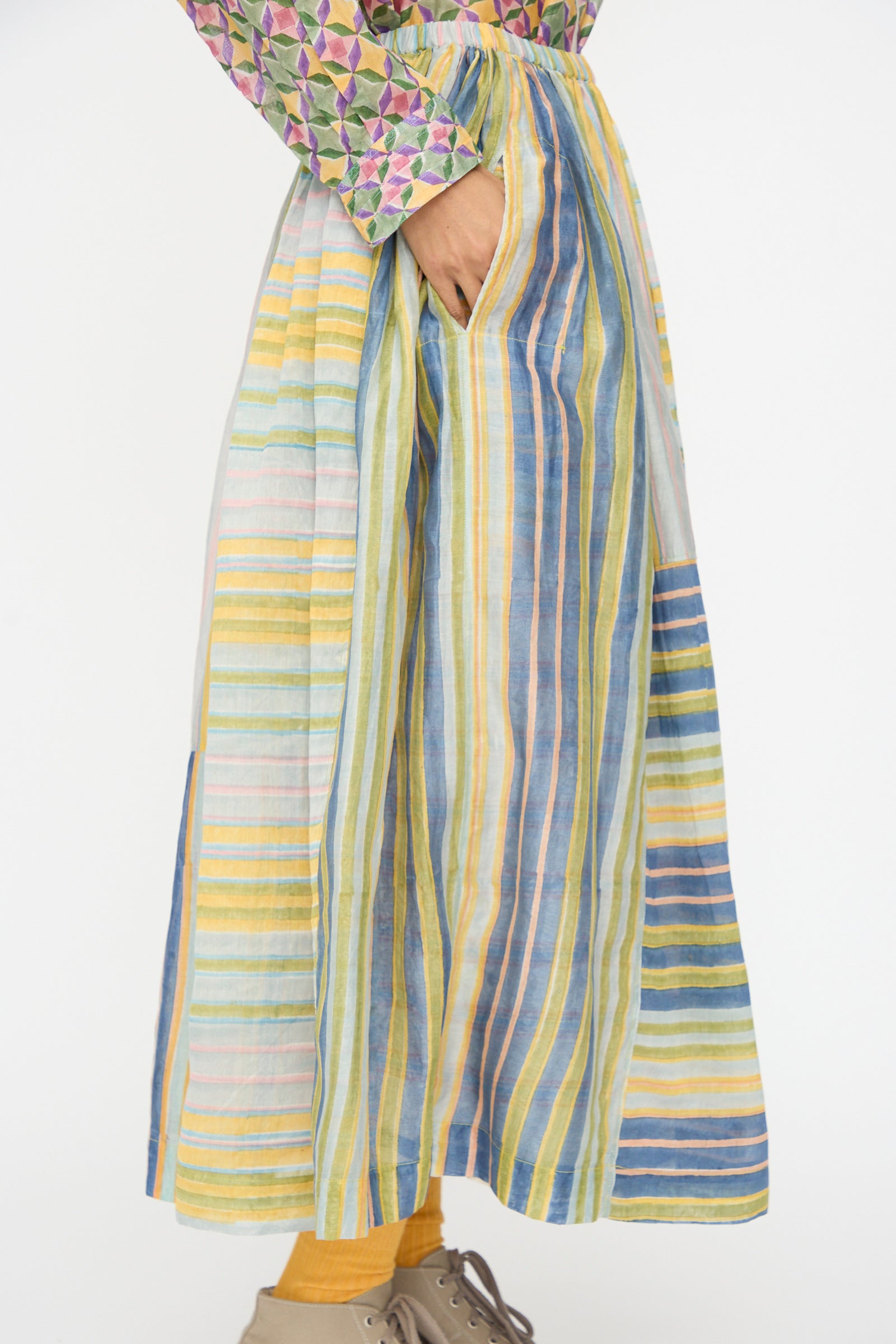 A person wears the Injiri Blockprint Chanderi Zamila Skirt in Green Multi with a hand in the pocket, paired with yellow tights, beige shoes, and a patterned long-sleeve top.