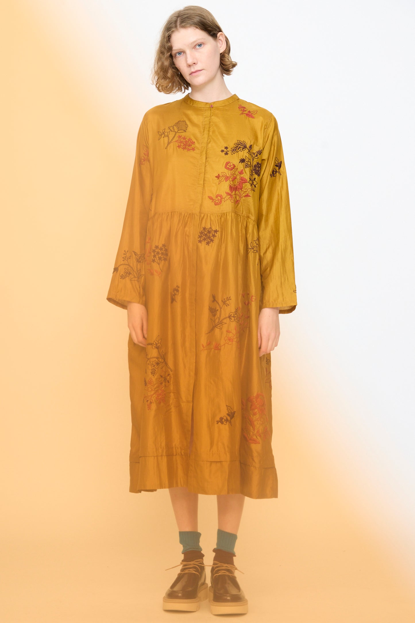 A person wearing the Injiri Chikankari Silk Naomi Dress in Ochre, featuring loose long sleeves and intricate floral embroidery, stands against a pale background with a yellow haze at the bottom.