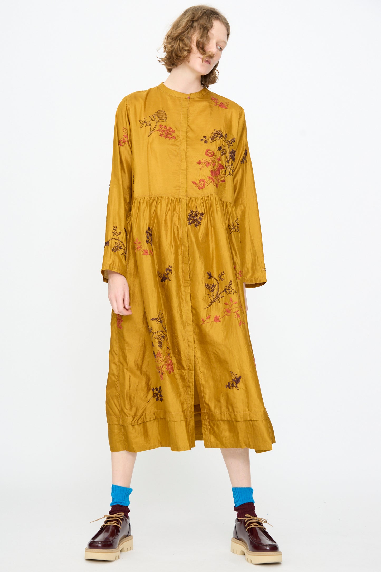 A person stands against a plain white background wearing the Injiri Chikankari Silk Naomi Dress in Ochre—an elegant loose-fitting, mustard yellow midi dress adorned with delicate floral embroidery—paired with blue socks and burgundy platform shoes.