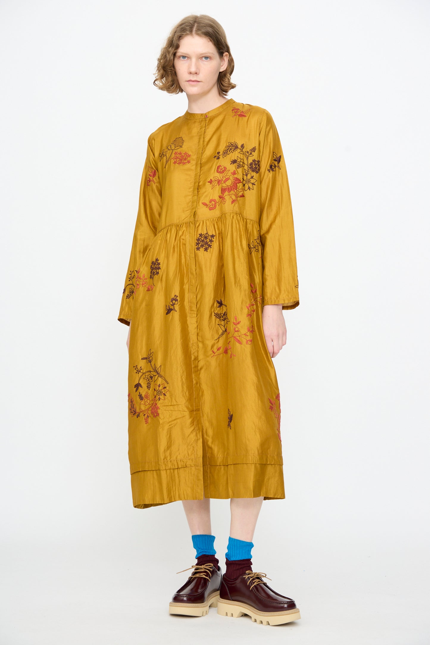 A person wearing the Injiri Chikankari Silk Naomi Dress in Ochre, featuring long sleeves and floral embroidery, paired with blue socks and brown platform shoes, stands against a plain white background.