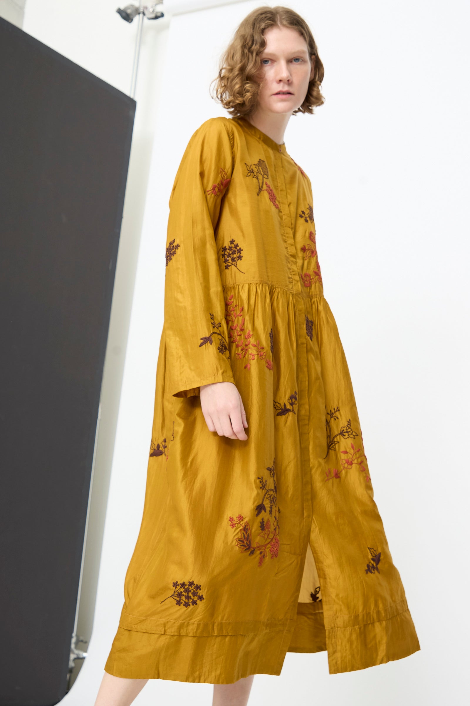 A person stands in a studio, facing slightly to the side, wearing the Injiri Chikankari Silk Naomi Dress in Ochre—a handcrafted silk dress featuring floral embroidery and long sleeves.