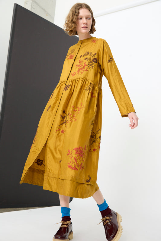 A person wearing the Injiri Chikankari Silk Naomi Dress in Ochre, a handcrafted silk dress featuring floral embroidery and long sleeves, paired with brown shoes and blue socks, walking in a minimalist indoor setting.