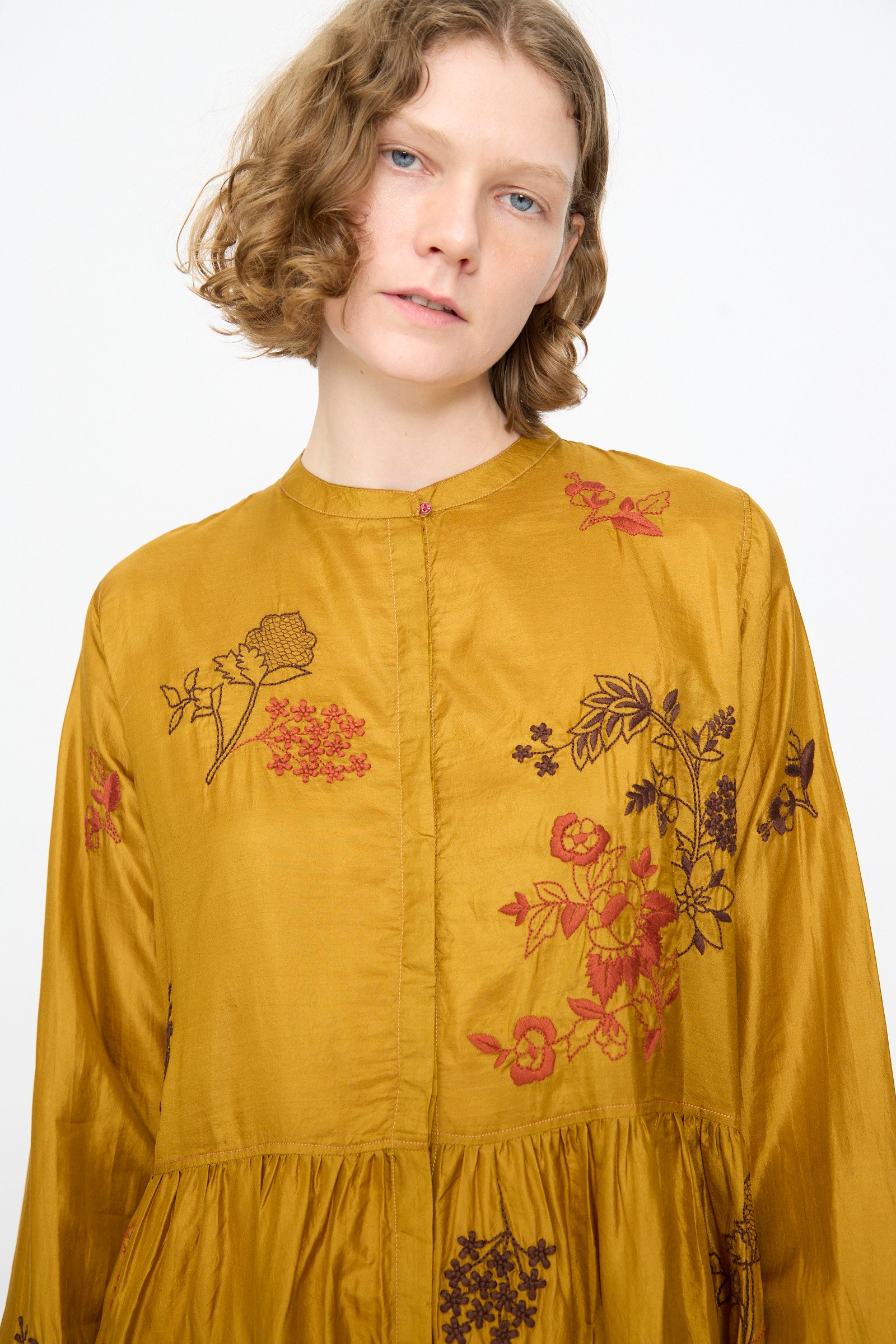 A person with short wavy hair wears the Injiri Chikankari Silk Naomi Dress in Ochre—handcrafted with elegant floral embroidery—standing against a plain white background.