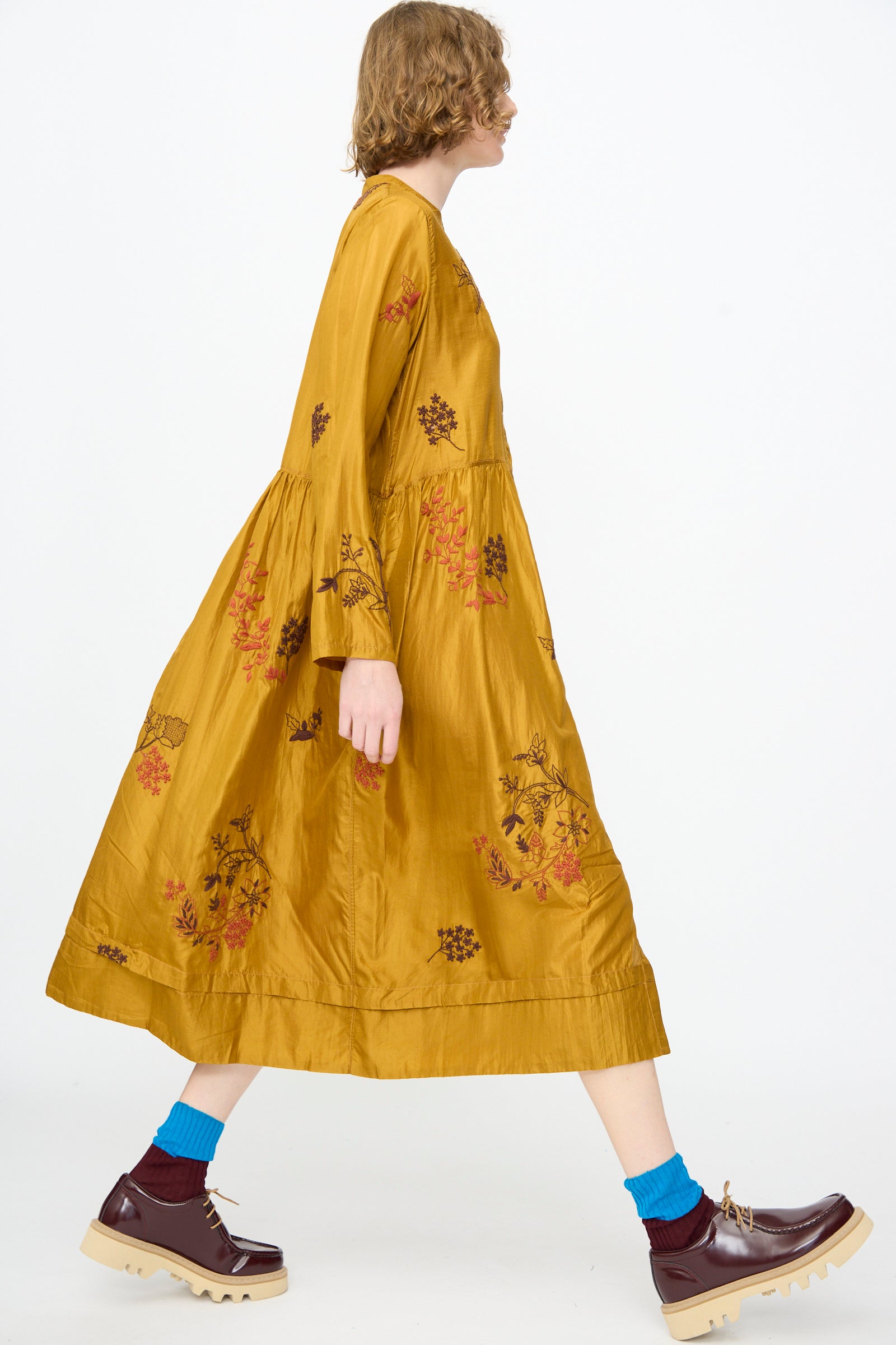 A person walks left against a plain white background, wearing the Injiri Chikankari Silk Naomi Dress in Ochre—an elegant, long-sleeved midi dress featuring intricate floral patterns—paired with blue socks and burgundy platform shoes.
