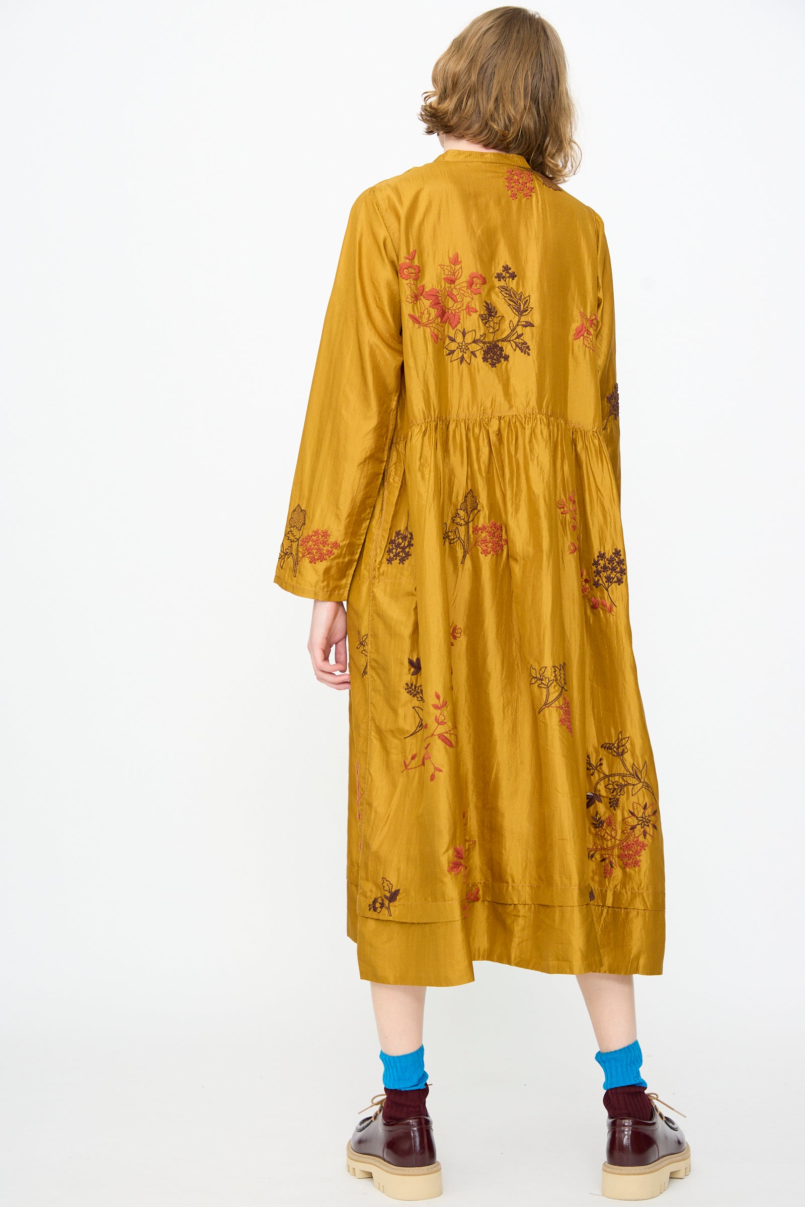 Wearing the Injiri Chikankari Silk Naomi Dress in Ochre, a handcrafted silk piece with long sleeves and delicate floral embroidery, the person is paired with maroon shoes and blue socks, standing with their back to the camera against a plain white background.