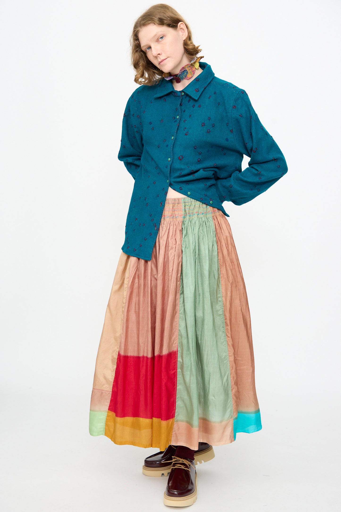 A person wearing a teal button-up shirt, the Injiri Clamp Dyed Silk Cotton Alisha Skirt in Multi featuring delicate hand embroidery, maroon platform shoes, and a floral neck scarf stands against a plain white background.